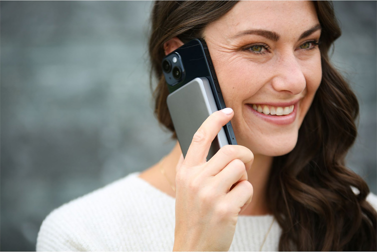 Frau telefoniert, hält Handy und graue Powerbank, lächelt.