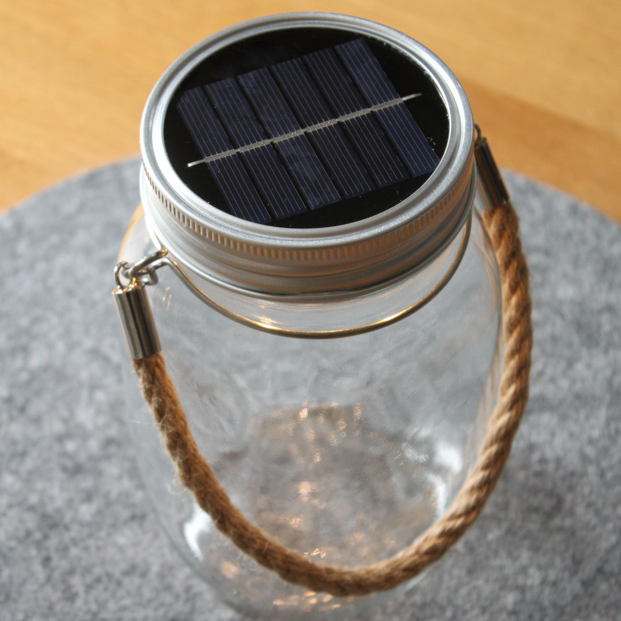 Glasbehälter mit Solarpanel oben, Seilgriff, auf grauer Oberfläche.