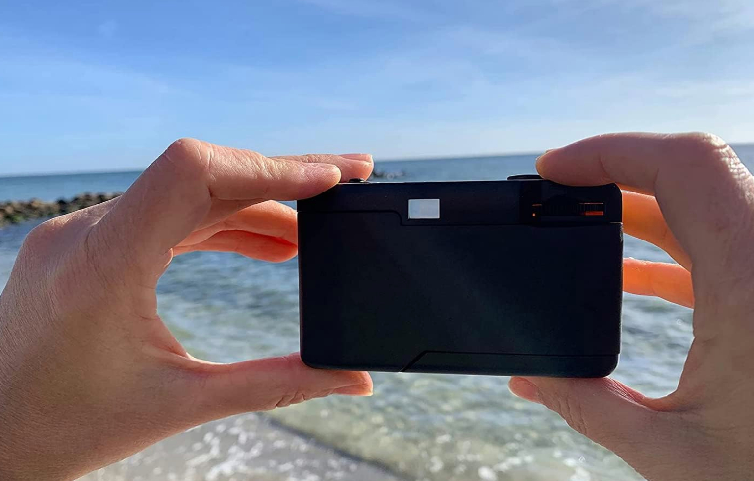 Eine Person hält eine schwarze Kamera vor dem Ozean und einem blauen Himmel.