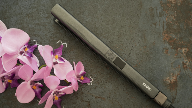 Gray hair straightener next to pink orchids on a dark, textured surface.