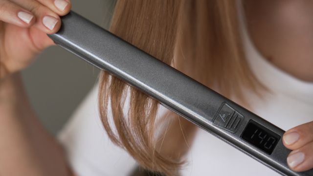 A person using a hair straightener on blonde hair, with the device showing the temperature.