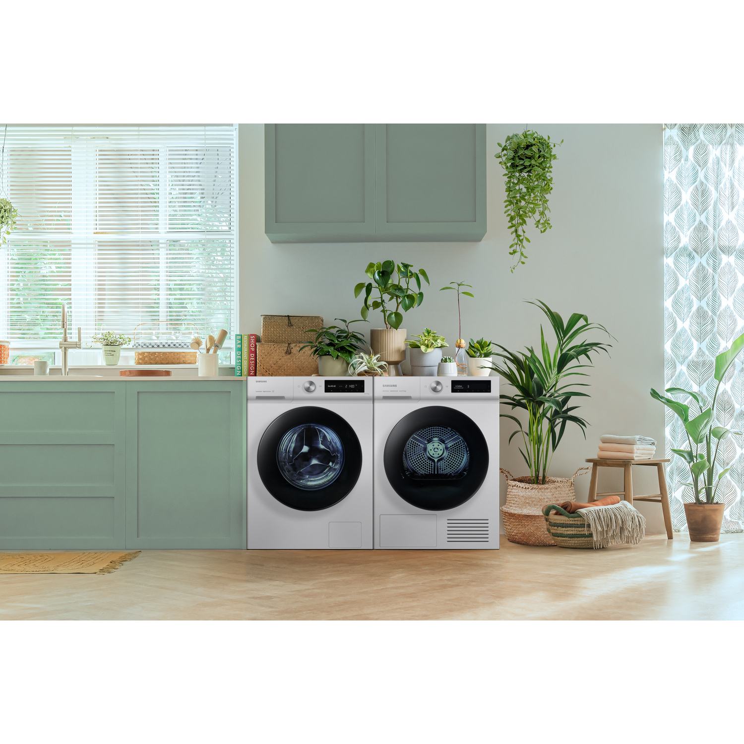 Une scène de cuisine avec un lave-linge et un sèche-linge. Il y a des plantes et de la décoration autour.