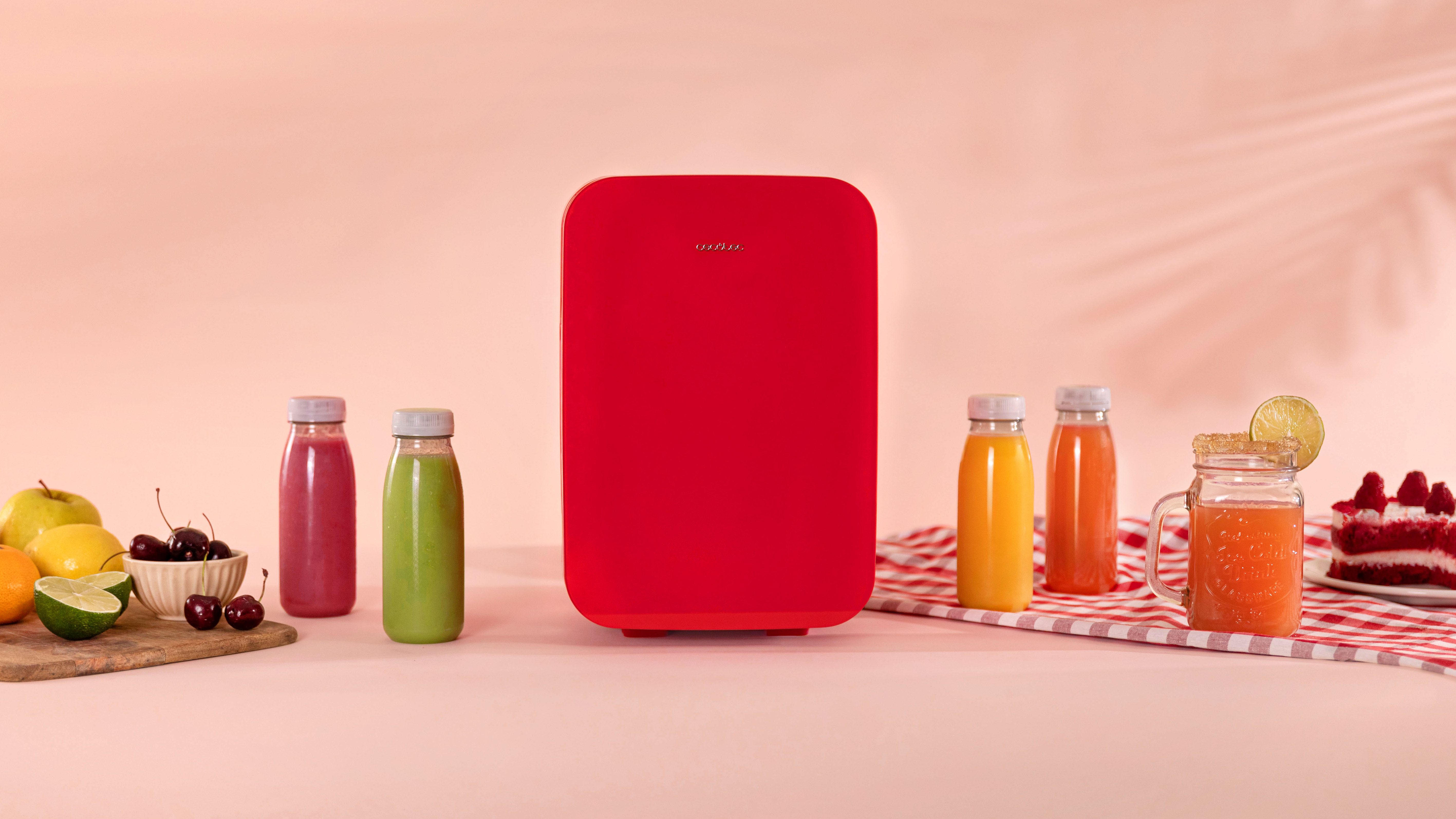 Una mini-nevera roja con botellas de zumo y fruta dispuestas sobre una superficie rosa. Hay un pastel.