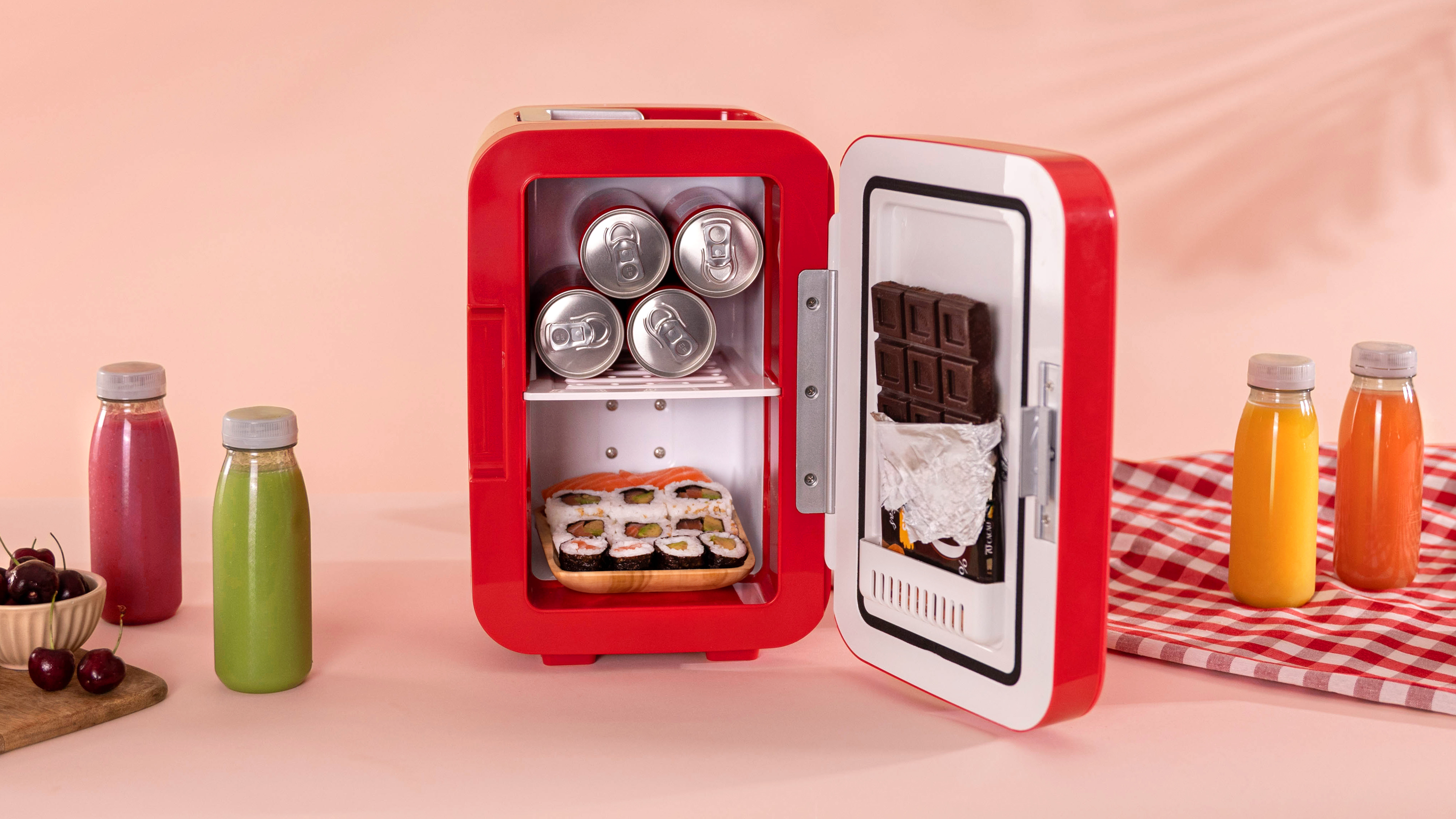 Una mini nevera roja con la puerta abierta. Latas, sushi y chocolate dentro. Botellas de zumo y cerezas cerca.