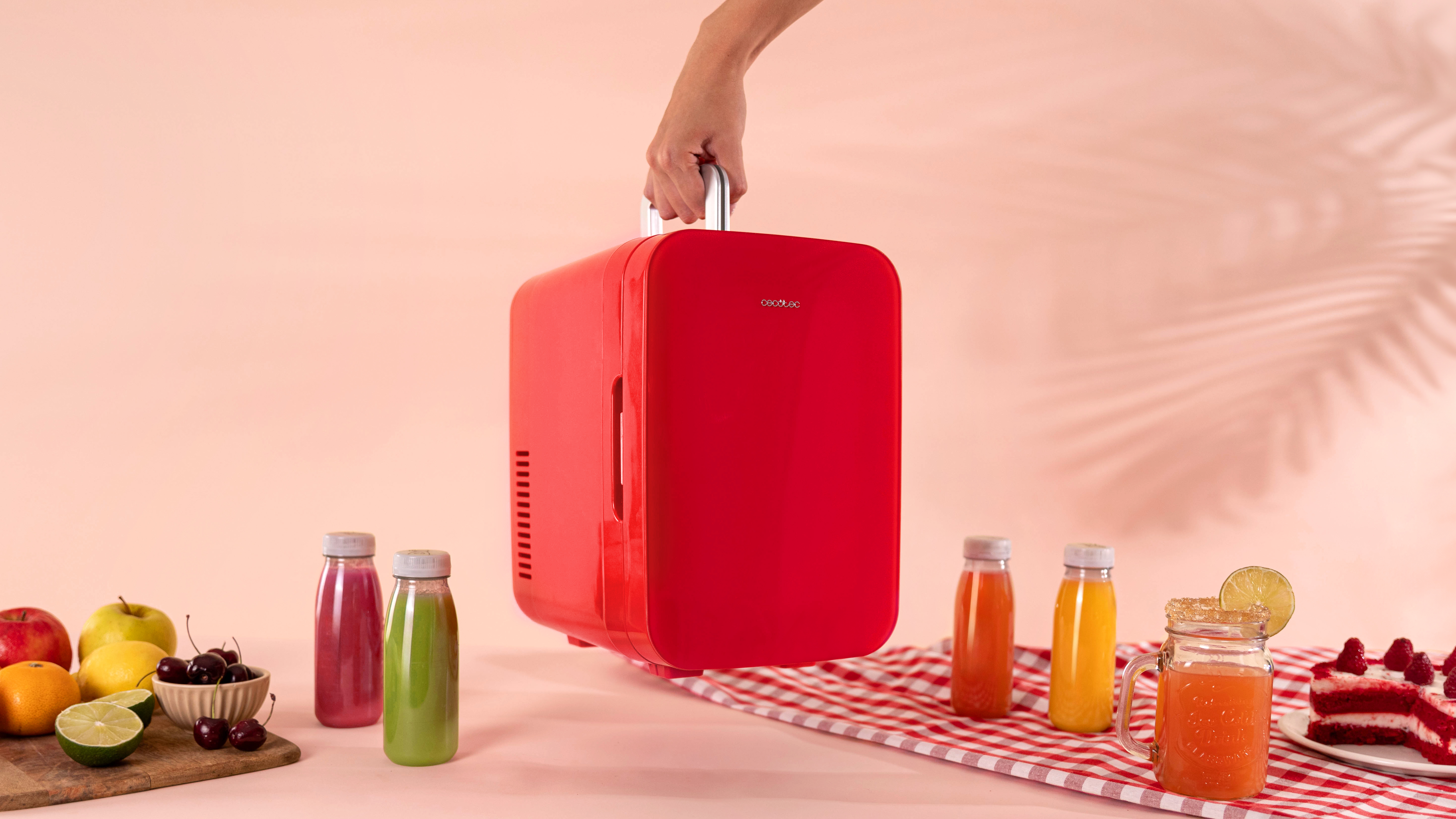 Mini nevera roja con asa, sostenida por una mano. Botellas y golosinas en la mesa.