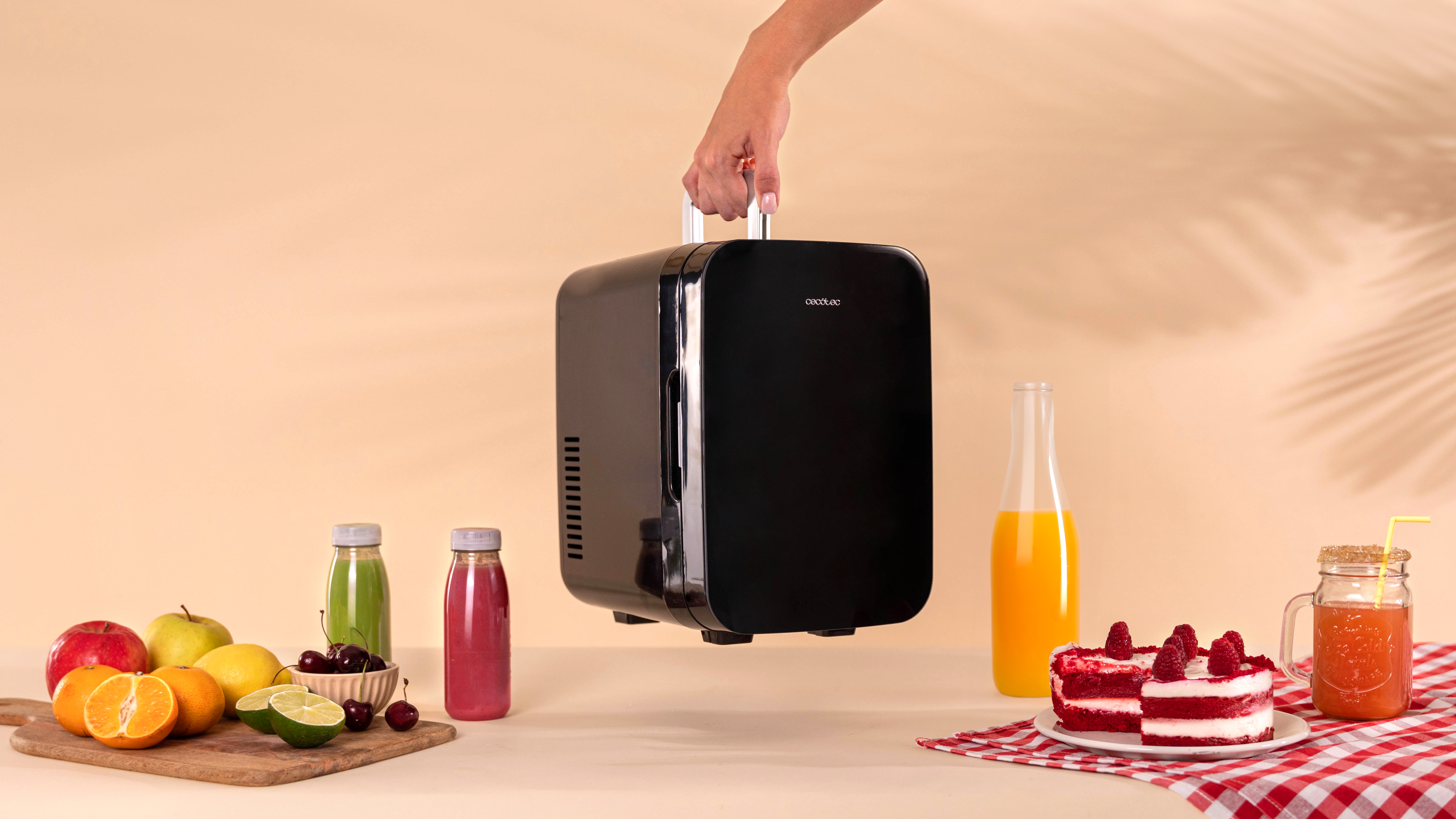 Mano sosteniendo un mini refrigerador negro. Frutas, zumos y pastel sobre la mesa.
