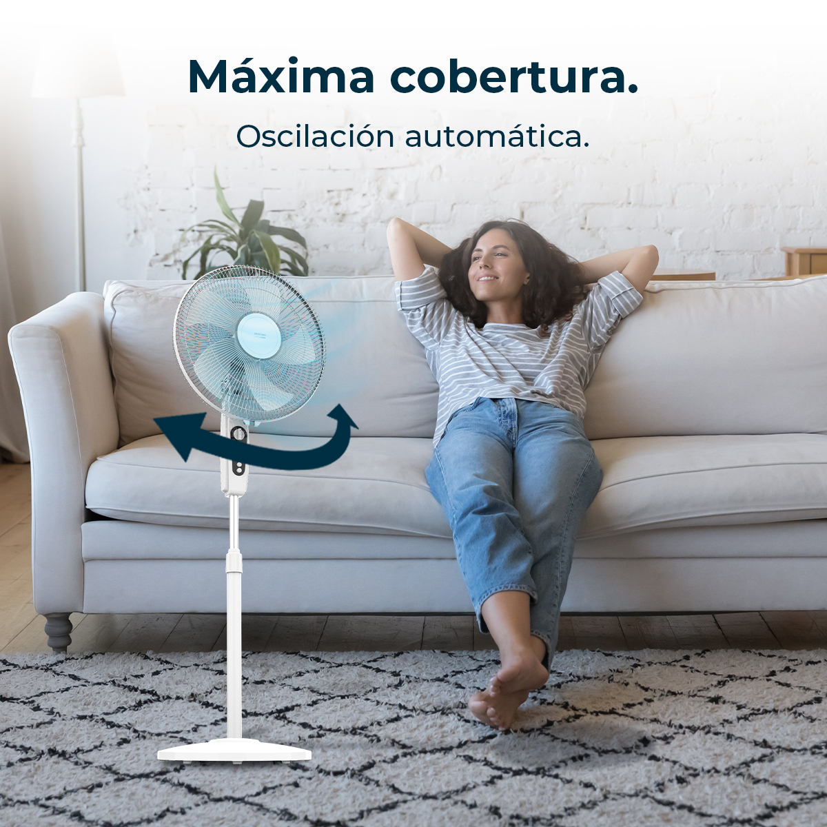 Una mujer se relaja cerca de un ventilador blanco. Está en un sofá blanco con una alfombra.