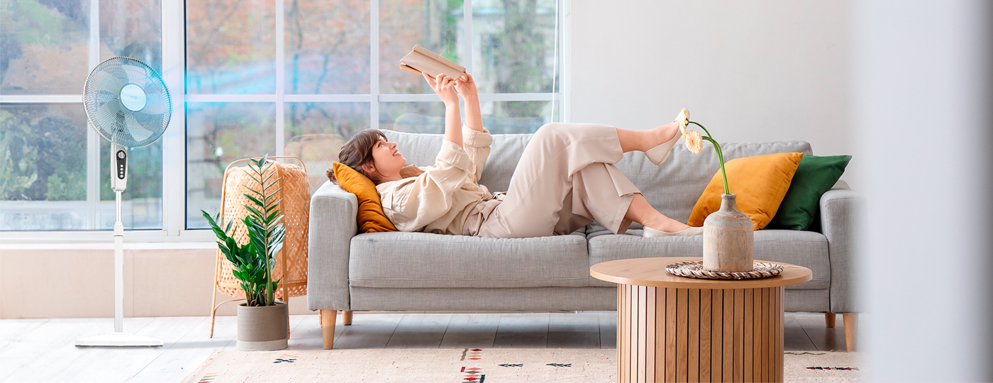 Mujer leyendo en sofá gris, con ventilador y planta al fondo. Mesa de madera cerca.