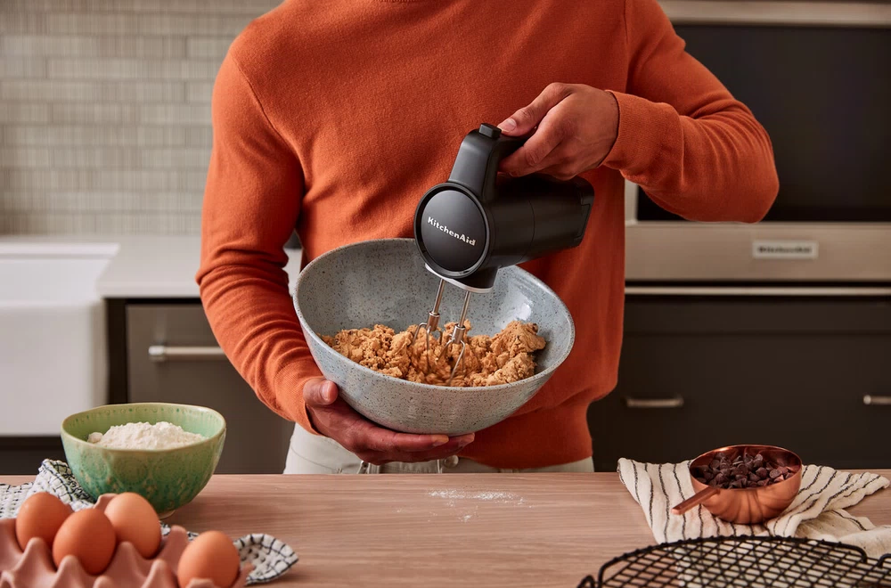 Narancssárga pulóveres személy, aki fekete KitchenAid keverővel keveri a süteménytésztát.