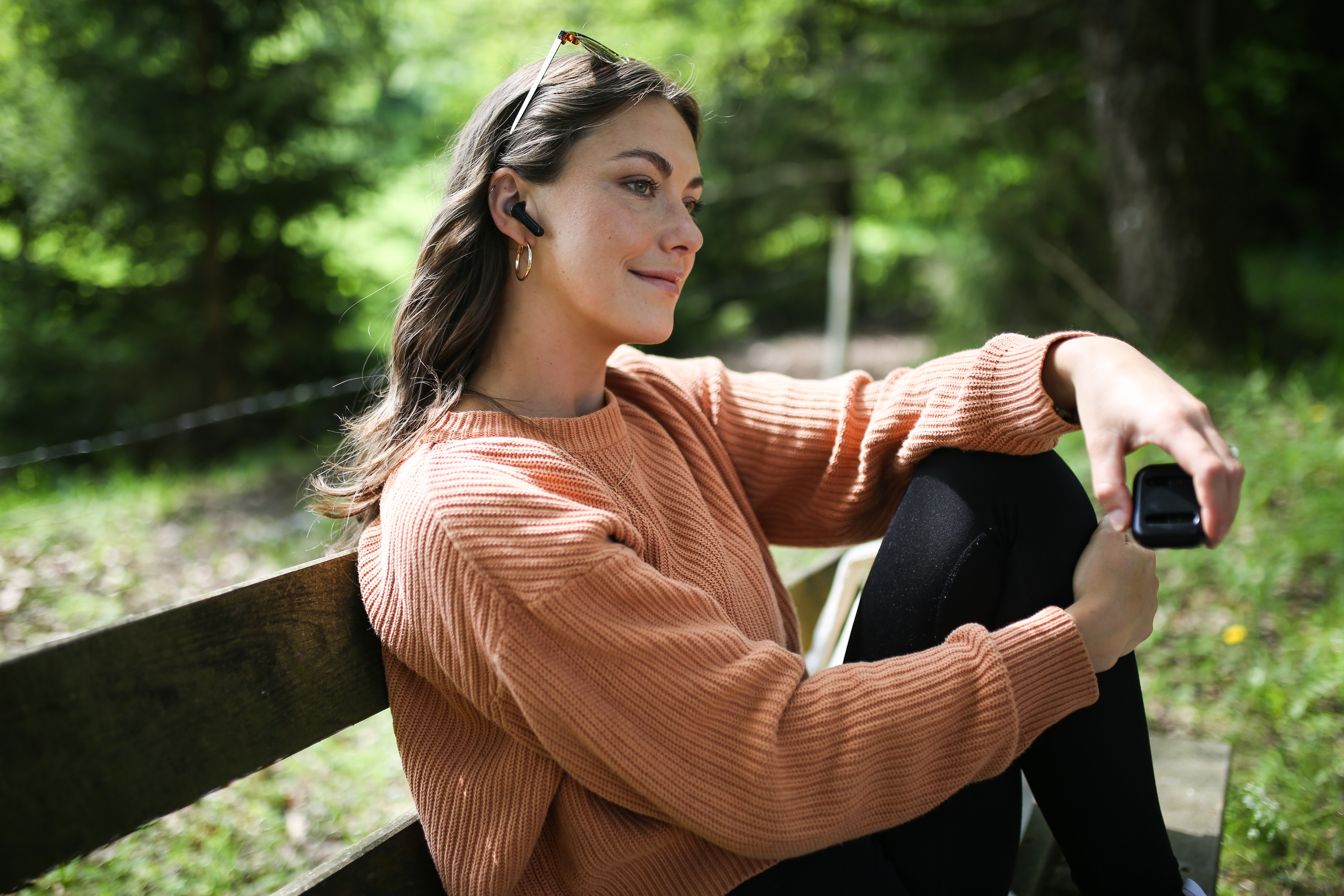 Kobieta siedzi na ławce w słuchawkach. Trzyma małe, czarne urządzenie. W tle drzewa.