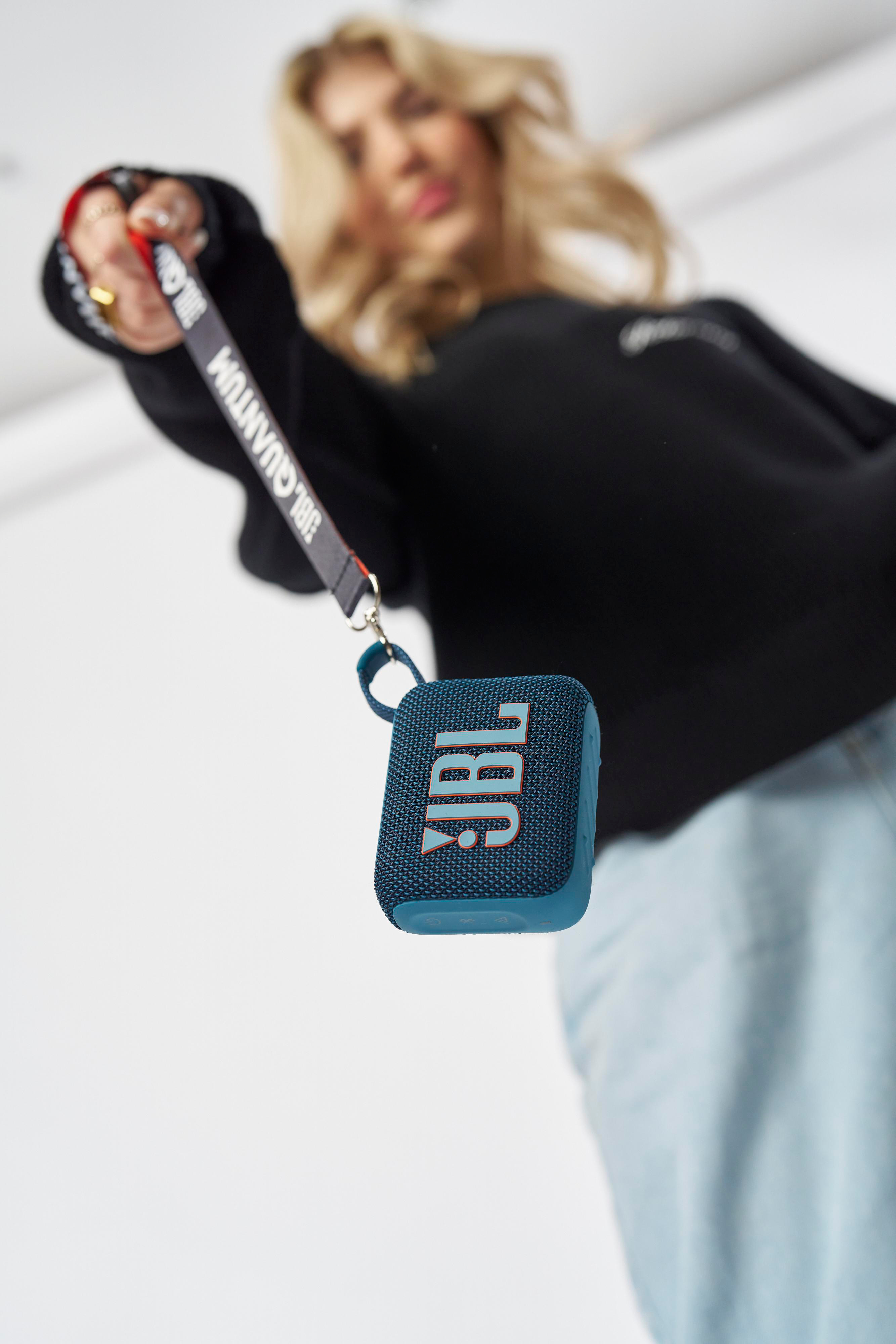 Eine Frau hält einen blauen JBL-Lautsprecher mit schwarzem Riemen. Der Hintergrund ist unscharf.