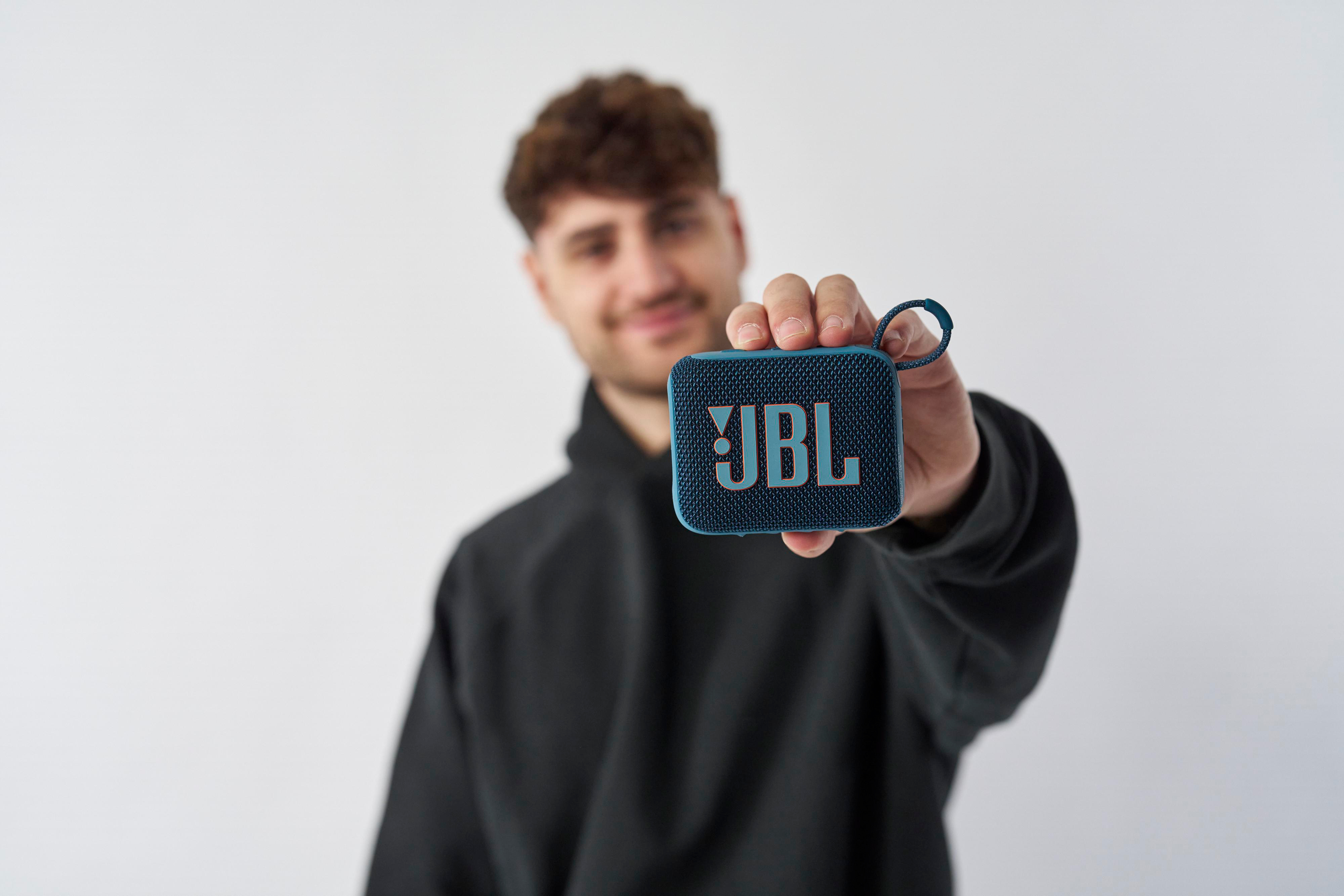 Ein Mann hält einen kleinen blauen JBL-Lautsprecher. Weißer Hintergrund, lächelnd.