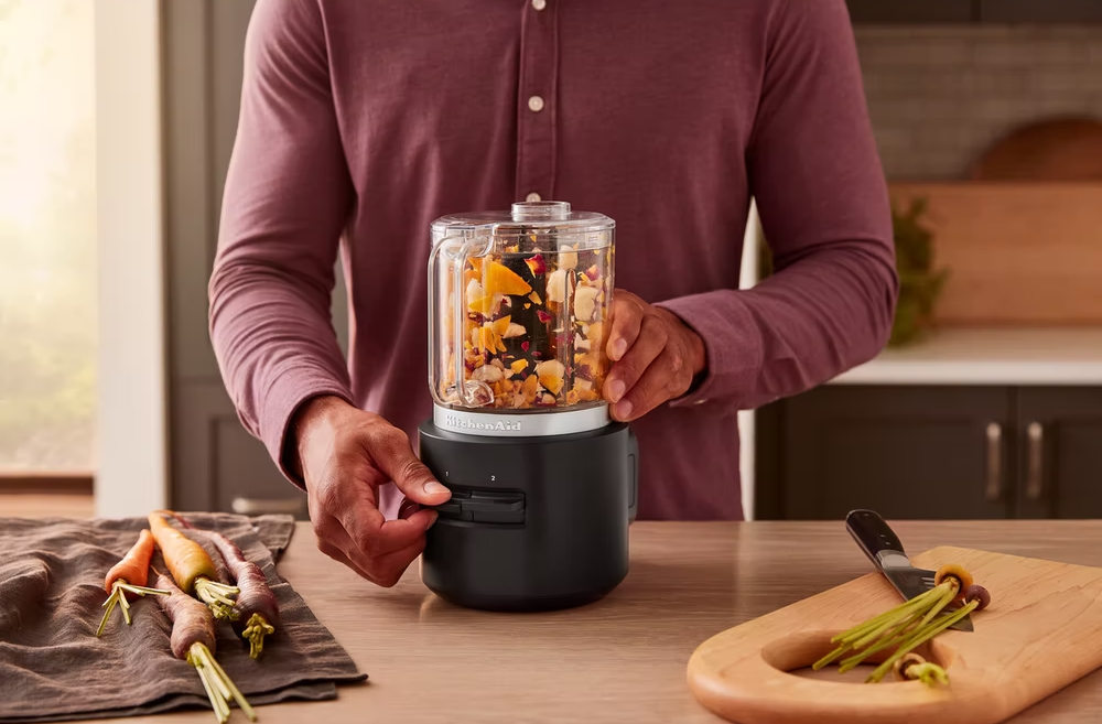 Egy személy KitchenAid konyhai robotgépet használ zöldségekkel egy fa deszkán.
