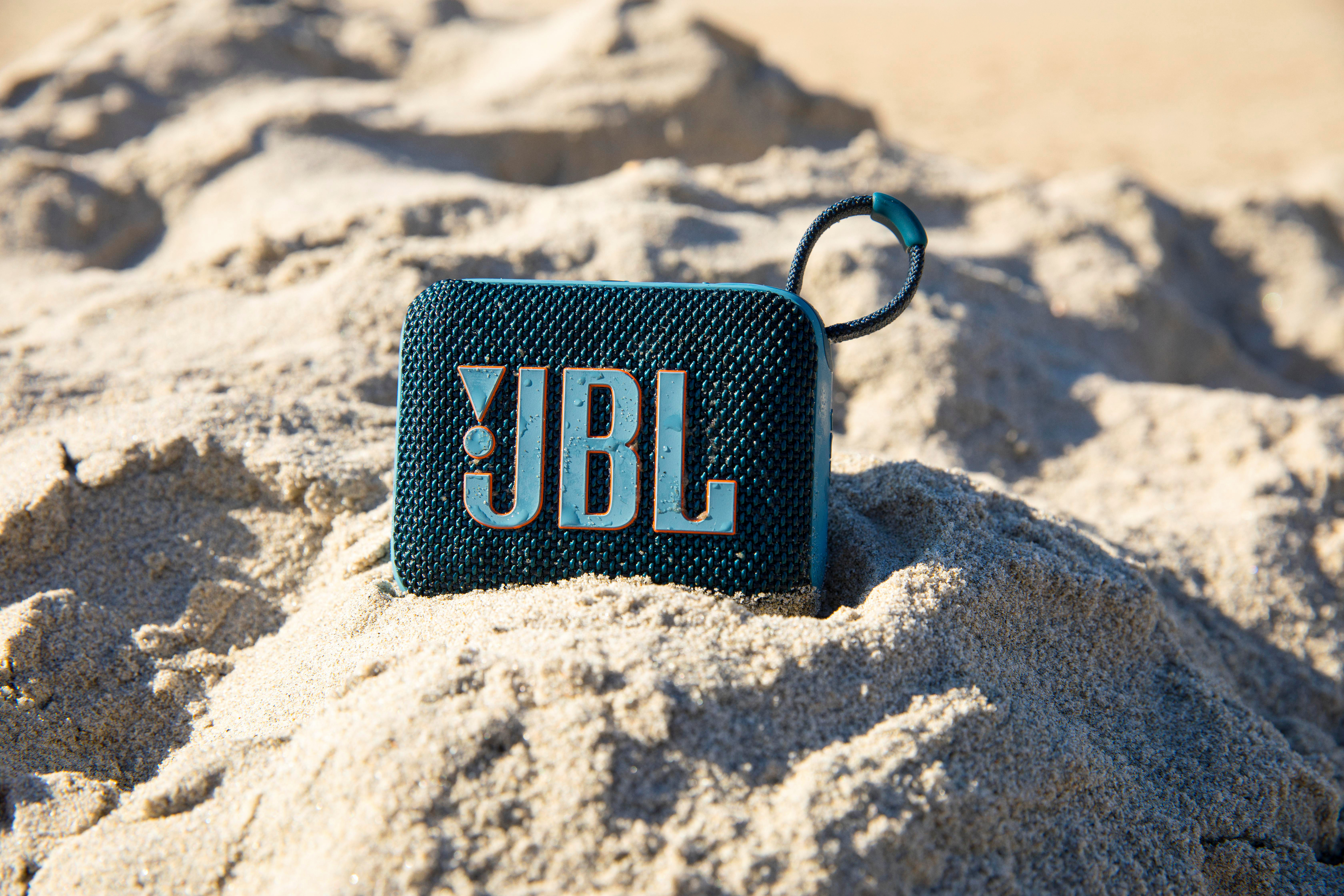 Ein kleiner, tealfarbener JBL-Lautsprecher auf Sand. Er hat einen schwarzen Griff und das JBL-Logo.