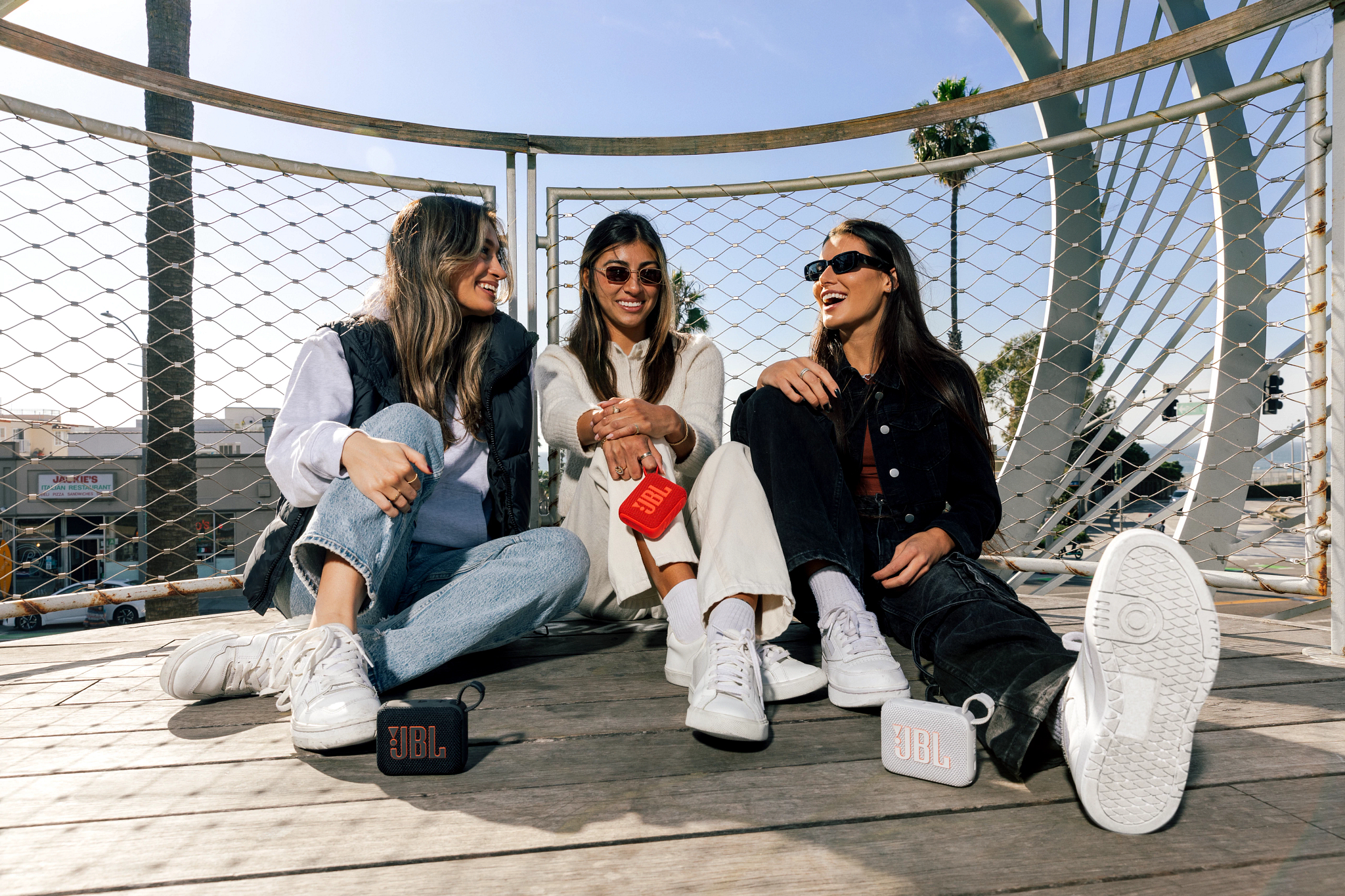 Drei Personen sitzen, lächeln, halten JBL-Lautsprecher auf einer Brücke.