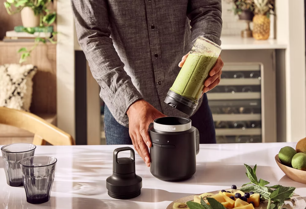 Egy személy egy turmixgép tartályát tartja. A turmixgép alapja az asztalon van, hozzávalókkal.