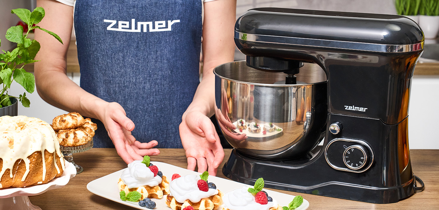 Kobieta w fartuchu stoi obok czarnego miksera stojącego z goframi i ciastem.