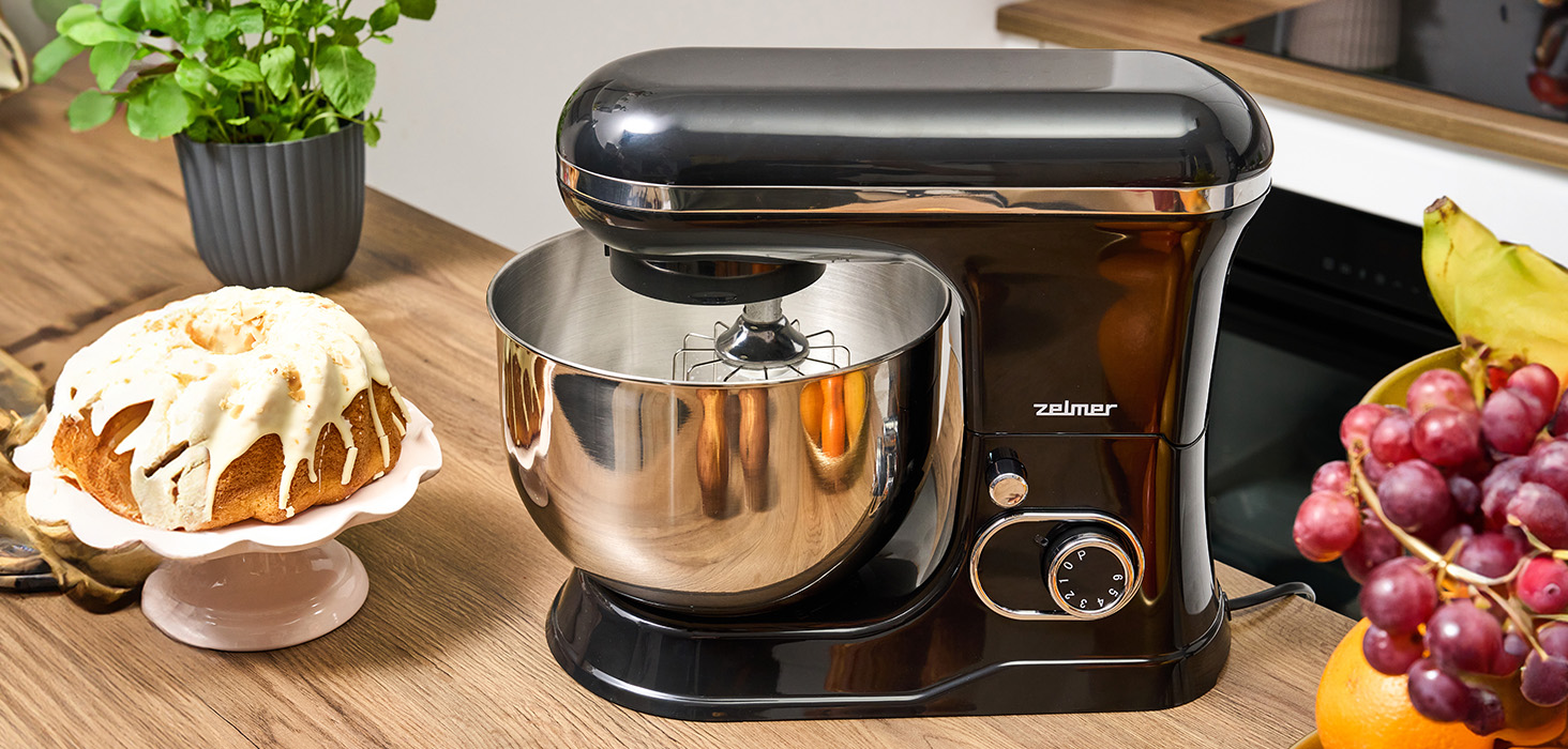 Czarny mikser stojący ze srebrną misą na drewnie. Ciasto i owoce w tle.