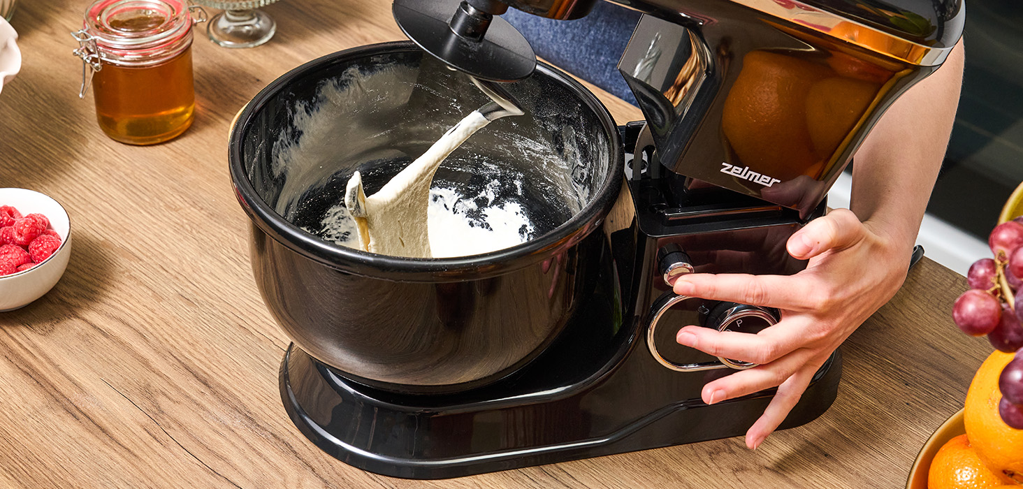 Osoba dodaje ciasto do czarnego miksera. Otoczenie kuchni z innymi składnikami.