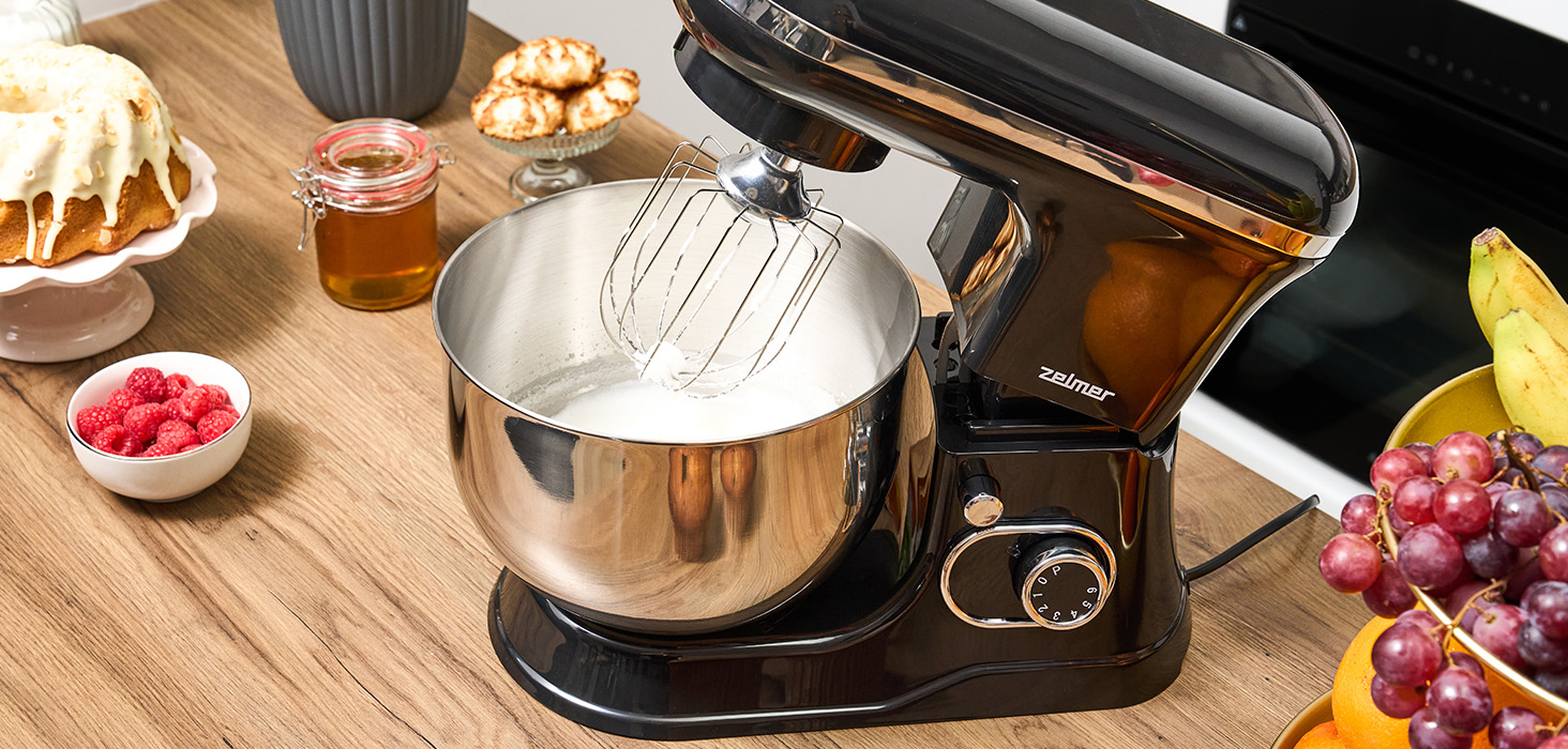 Czarny mikser stojący z miską z białą mieszanką. Blat kuchenny z produktami spożywczymi.