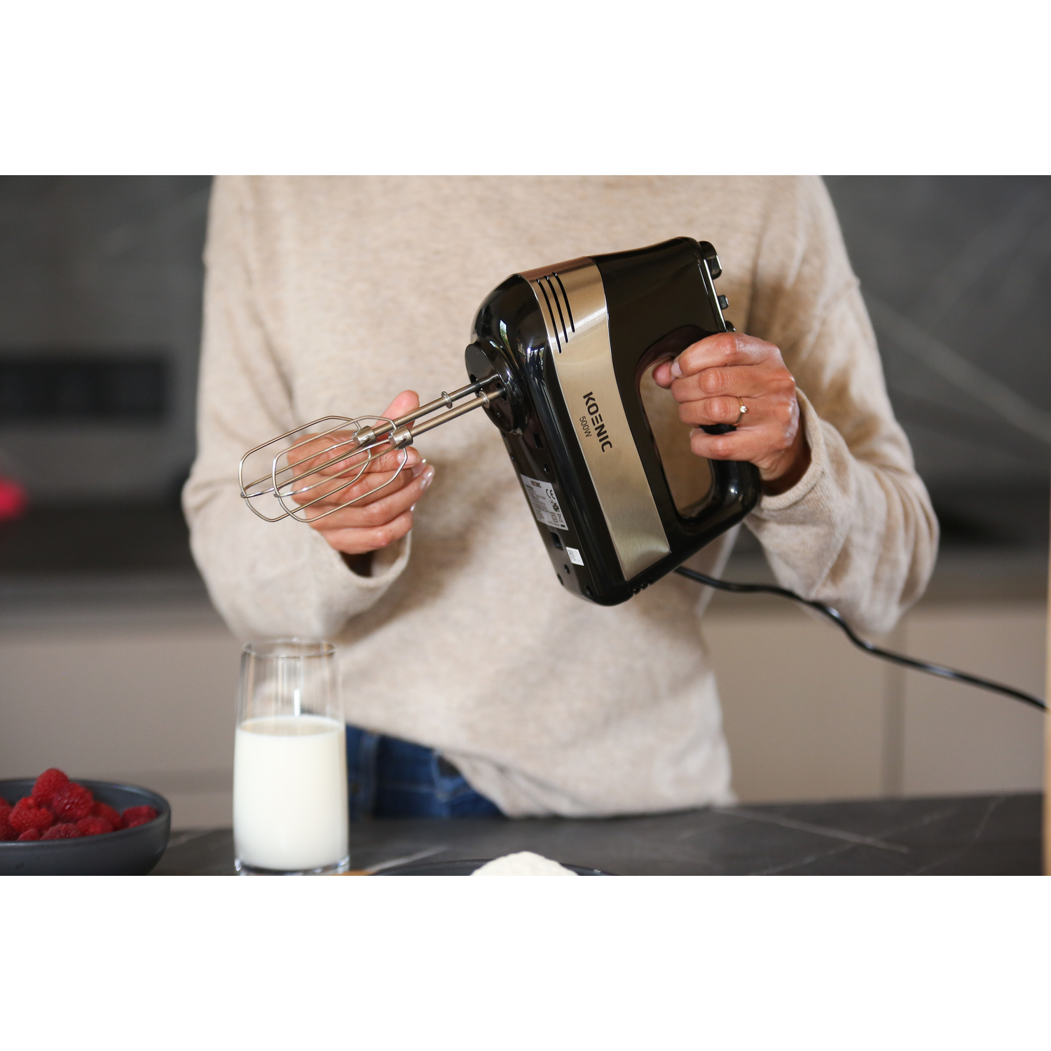 A person holding a black and silver hand mixer. A glass of milk and raspberries are on the table.