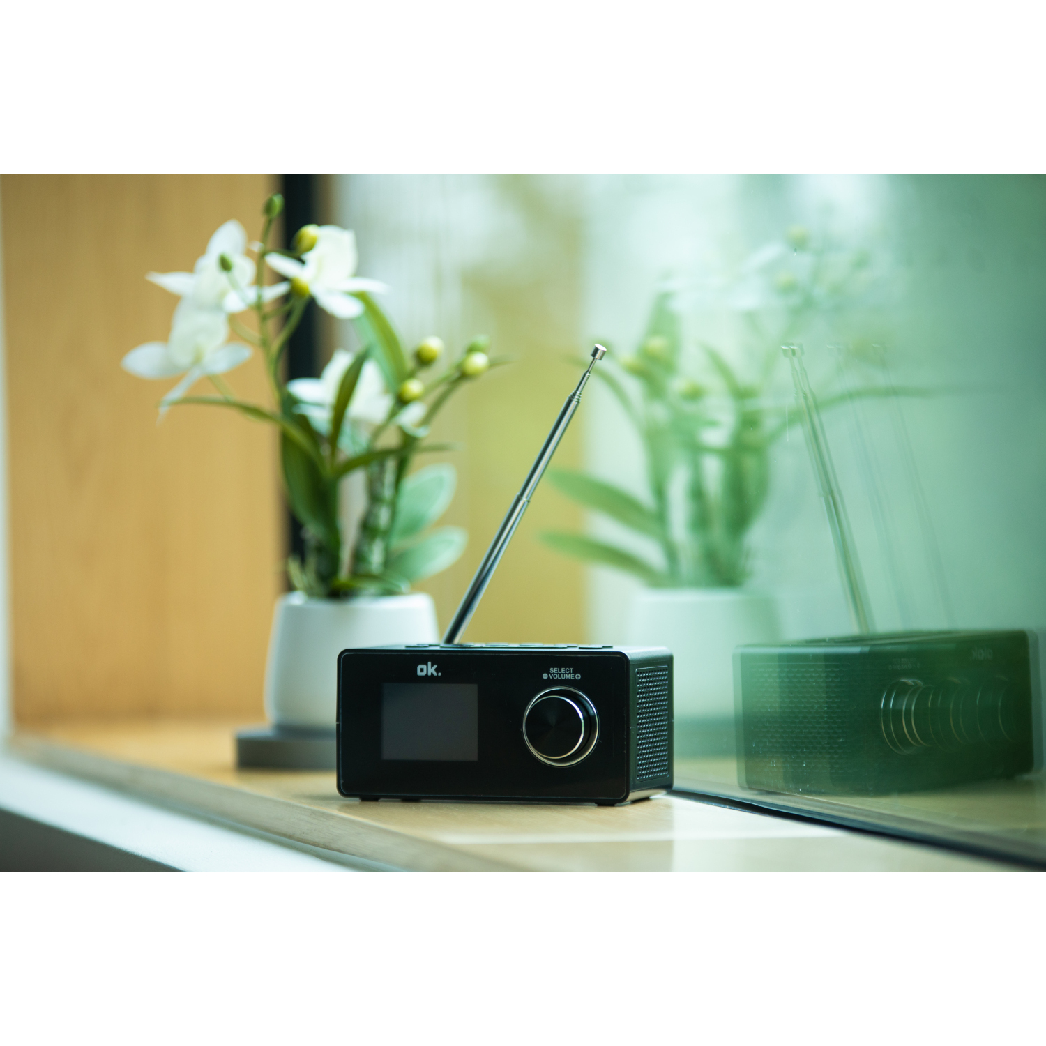 Een zwarte radio met zilveren antenne staat op een vensterbank. Witte bloemen in potten staan op de achtergrond.