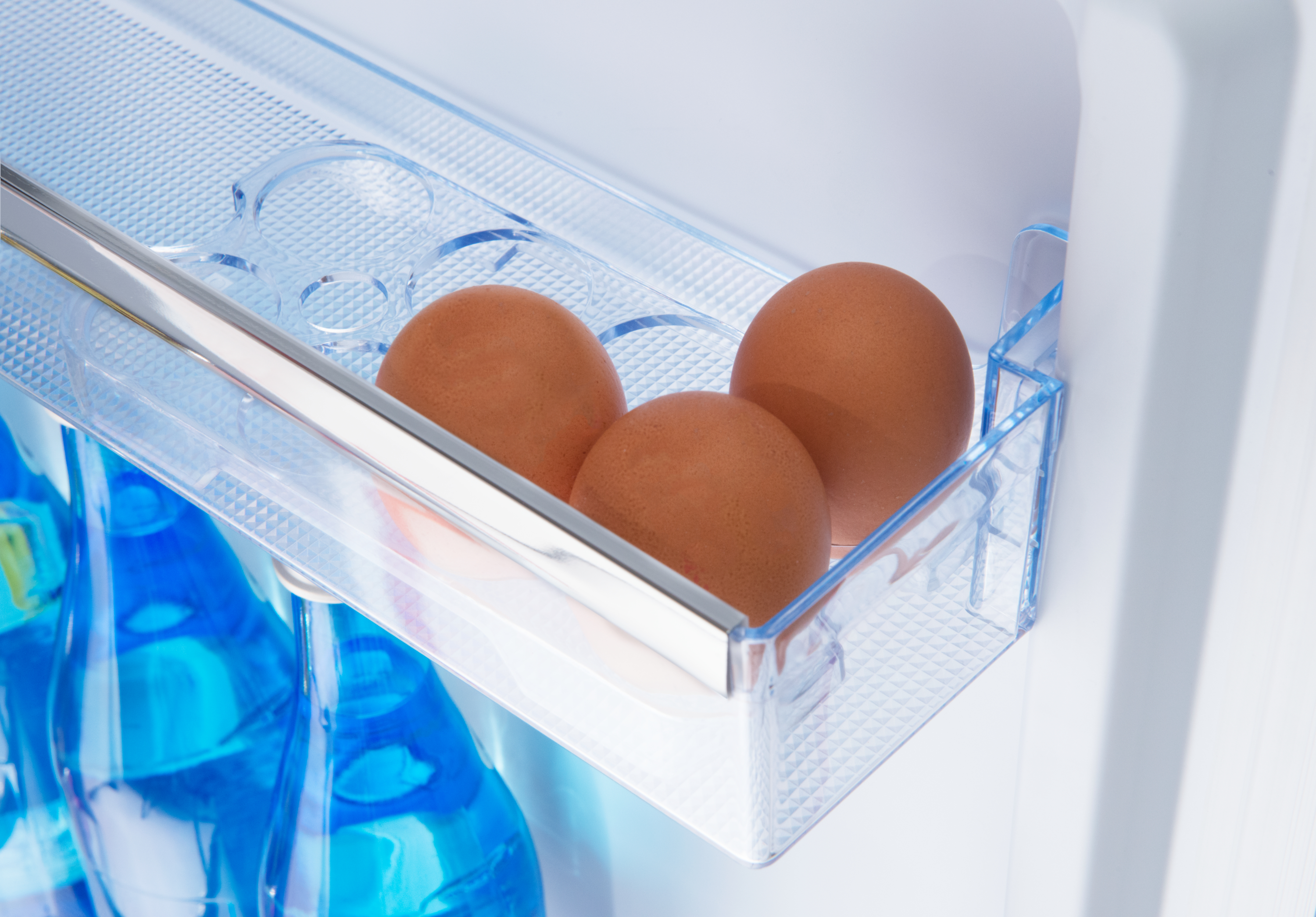 Drei braune Eier in einem durchsichtigen Kunststoffbehälter in einem Kühlschrank, mit blauen Flaschen im Hintergrund.
