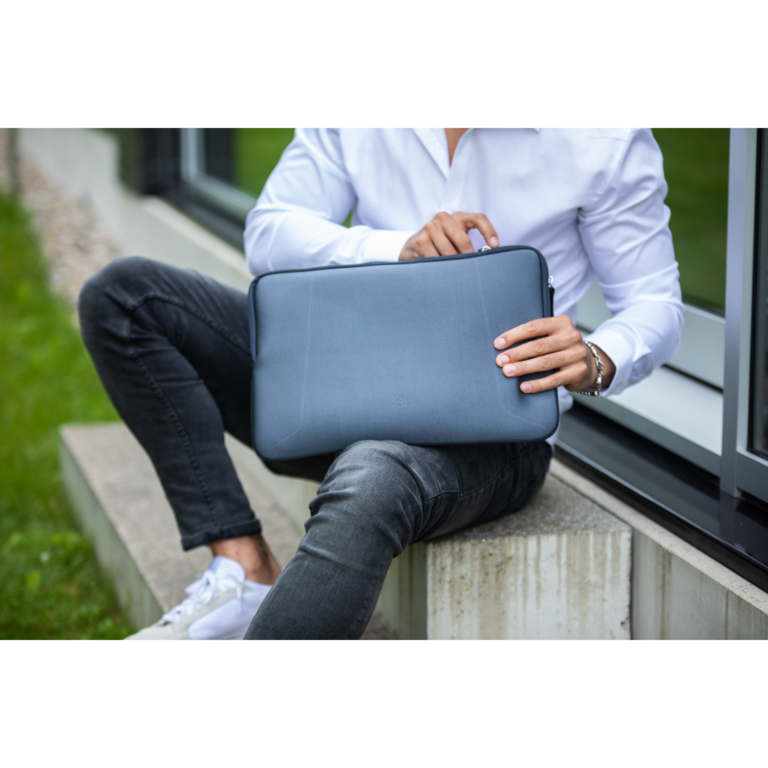 Man seated, holding a gray laptop case. He's wearing a white shirt and gray jeans.