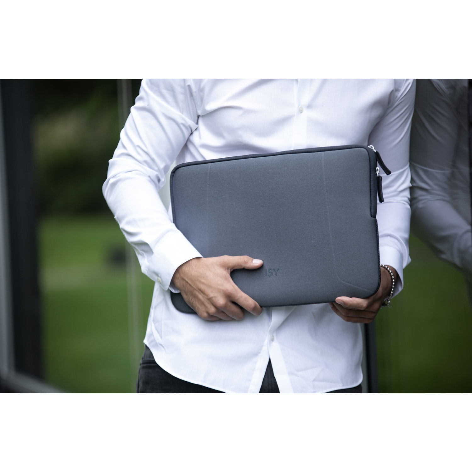 Man holding a gray laptop sleeve. White shirt and dark pants. Background of green plants and bright light.
