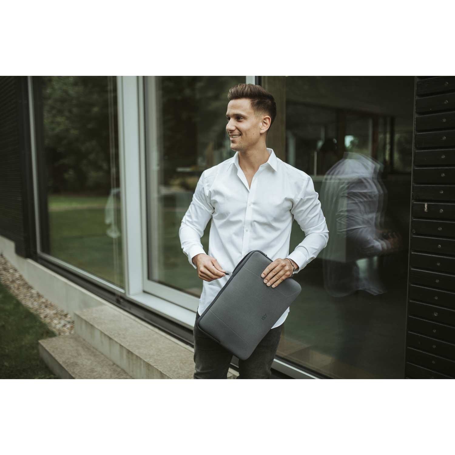 A man in a white shirt holding a gray laptop sleeve, smiling, outdoors.