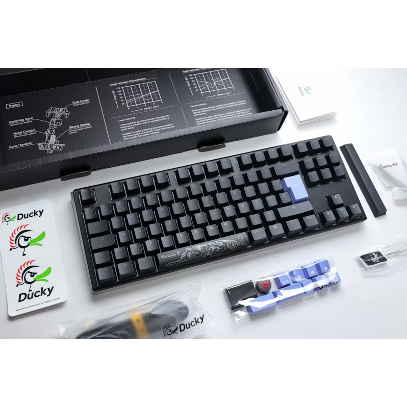Teclado negro con detalles azules. Logotipo de Ducky. Accesorios dispersos alrededor del teclado.