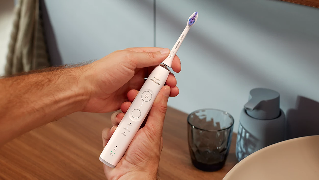 Kezek egy fehér elektromos fogkefét tartanak egy mosdókagyló közelében.