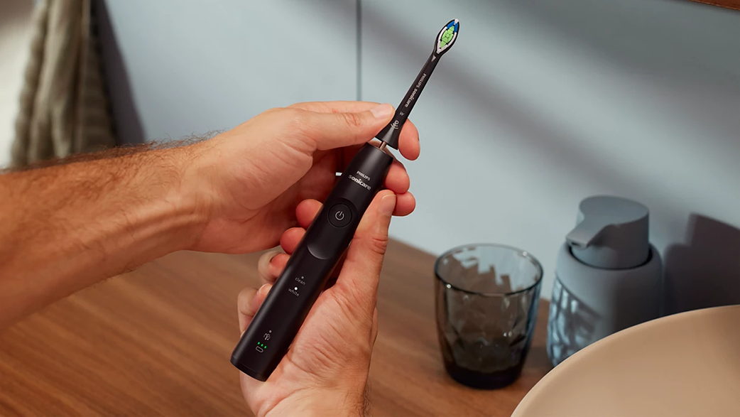 Kezek, amik egy fekete elektromos fogkefét tartanak. A feje zöld és kék.