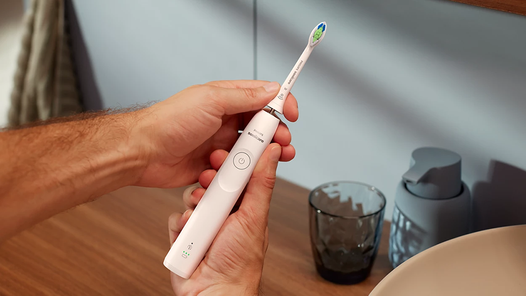 Kezek egy rózsaszín elektromos fogkefét tartanak. A háttérben egy mosdó és egy tükör látható.