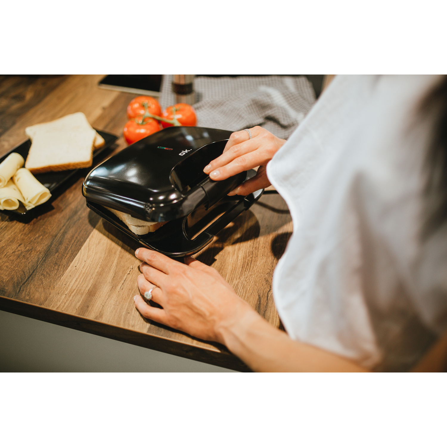 Een vrouw gebruikt een zwarte sandwichmaker op een houten aanrecht. Brood, tomaten en kaas zijn in de buurt.