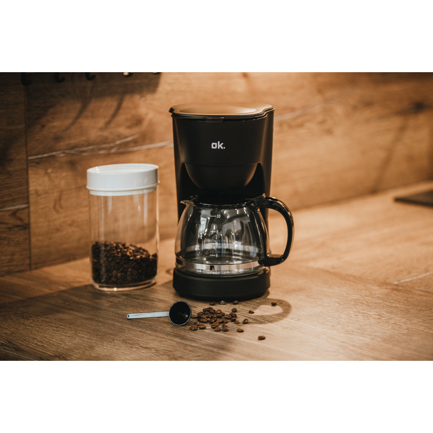 A coffee maker with a jar of beans and a spoon on a wooden table.