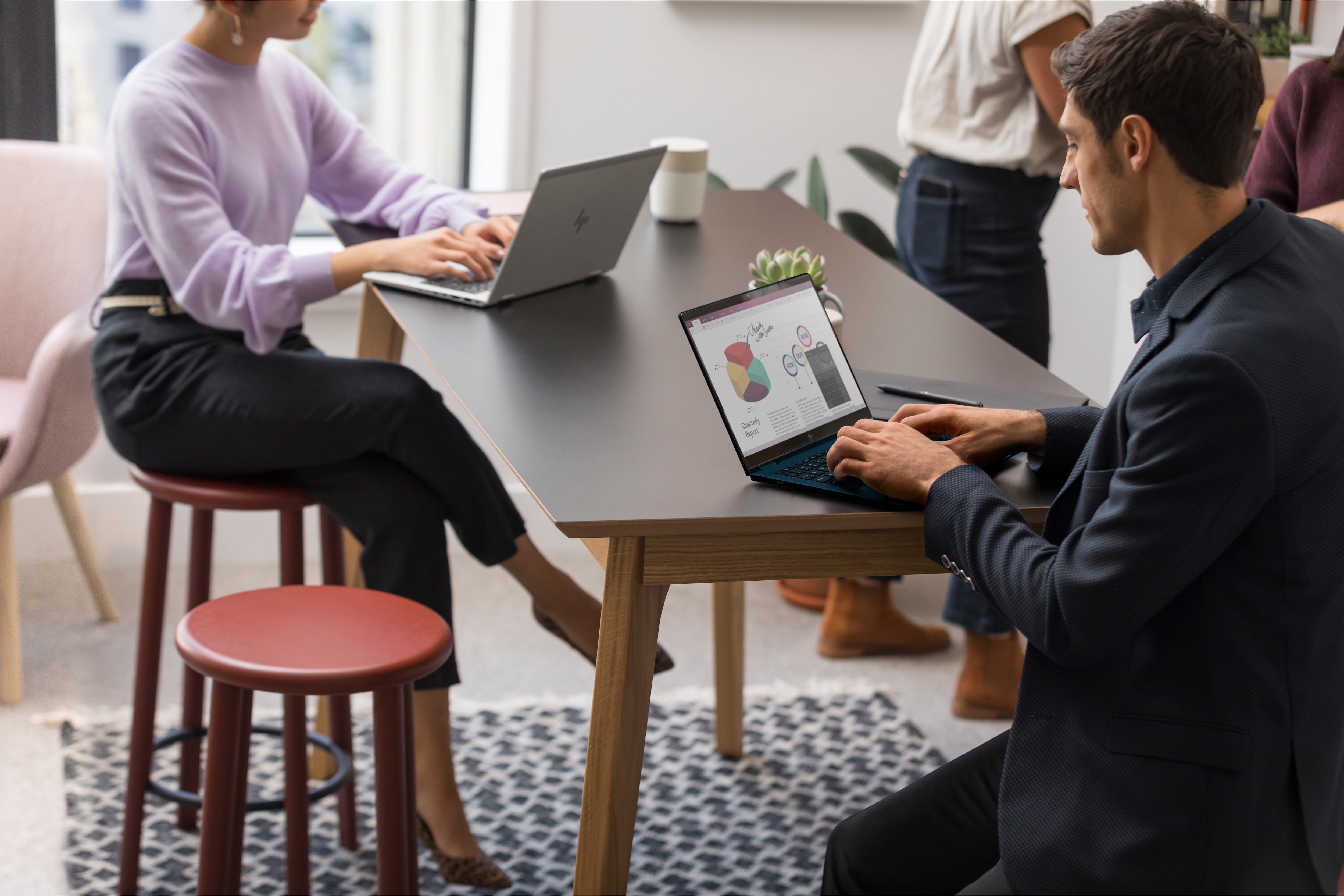 Leute arbeiten an einem Tisch mit Laptops. Eine Person sitzt auf einem roten Hocker. Helles und modernes Büro.