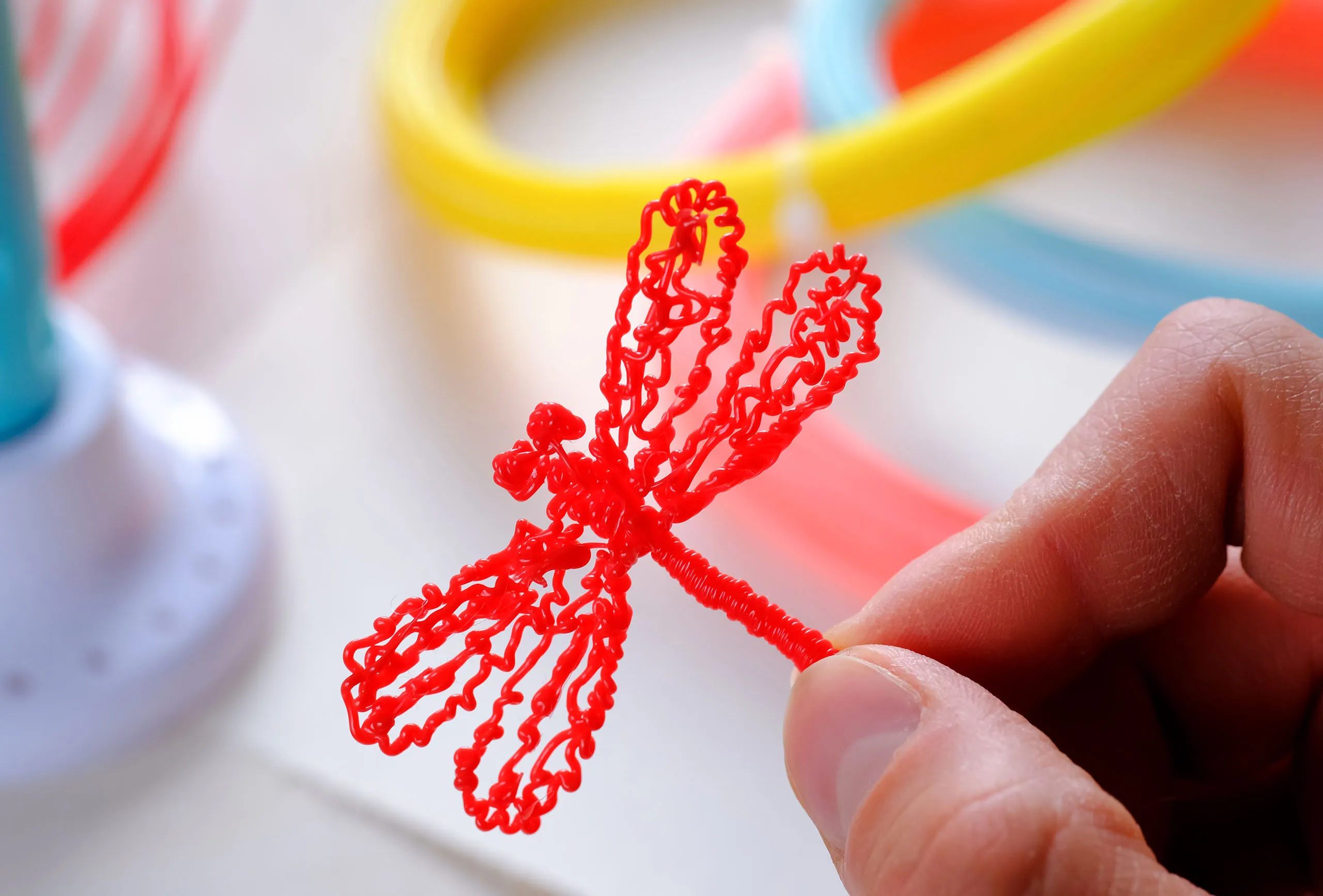 Libellule rouge fabriquée avec un stylo 3D. Tenue par une main, autres fils plastiques colorés.