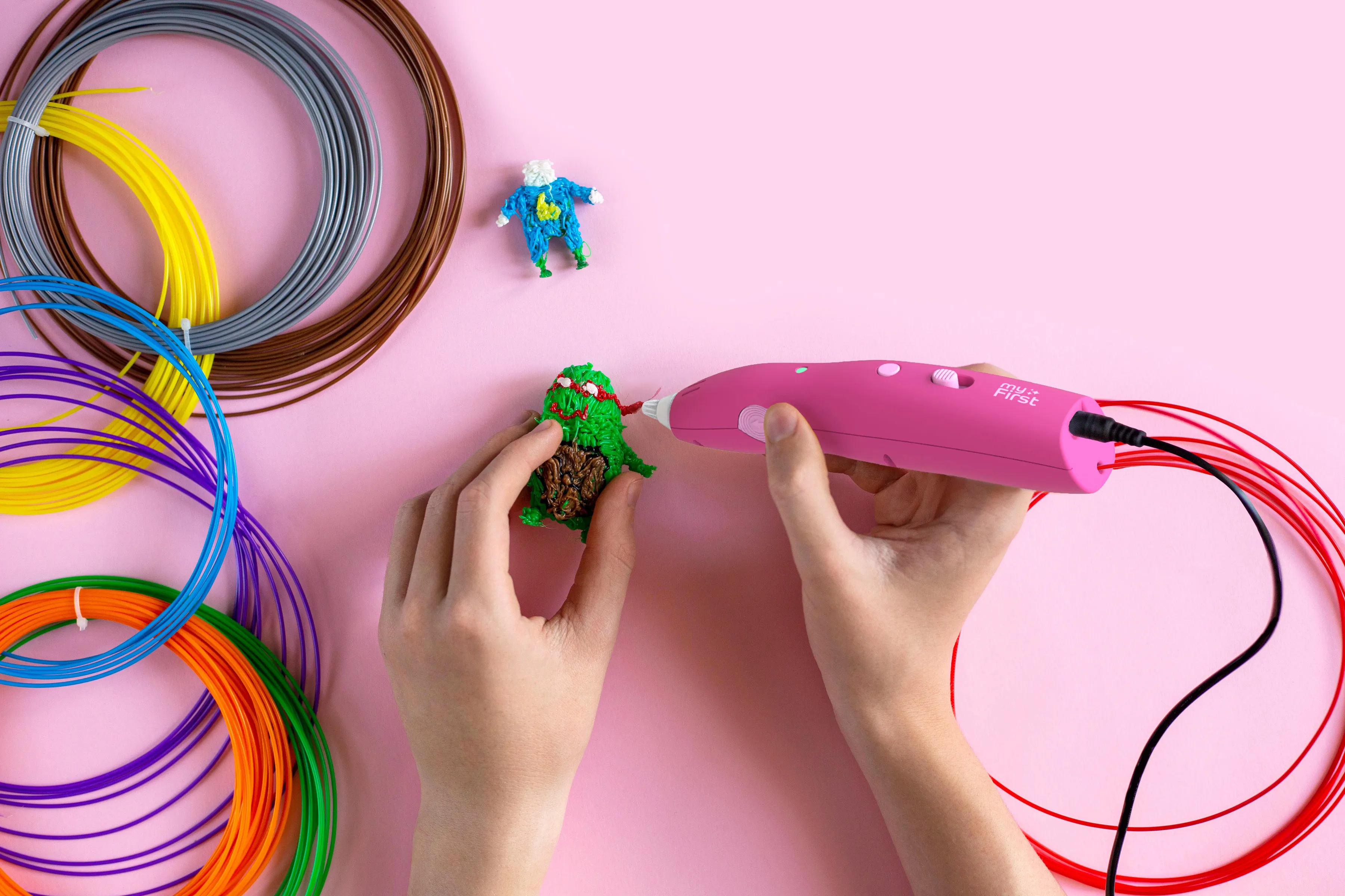 Mains tenant un modèle imprimé en 3D avec stylo rose et filaments colorés sur surface rose.