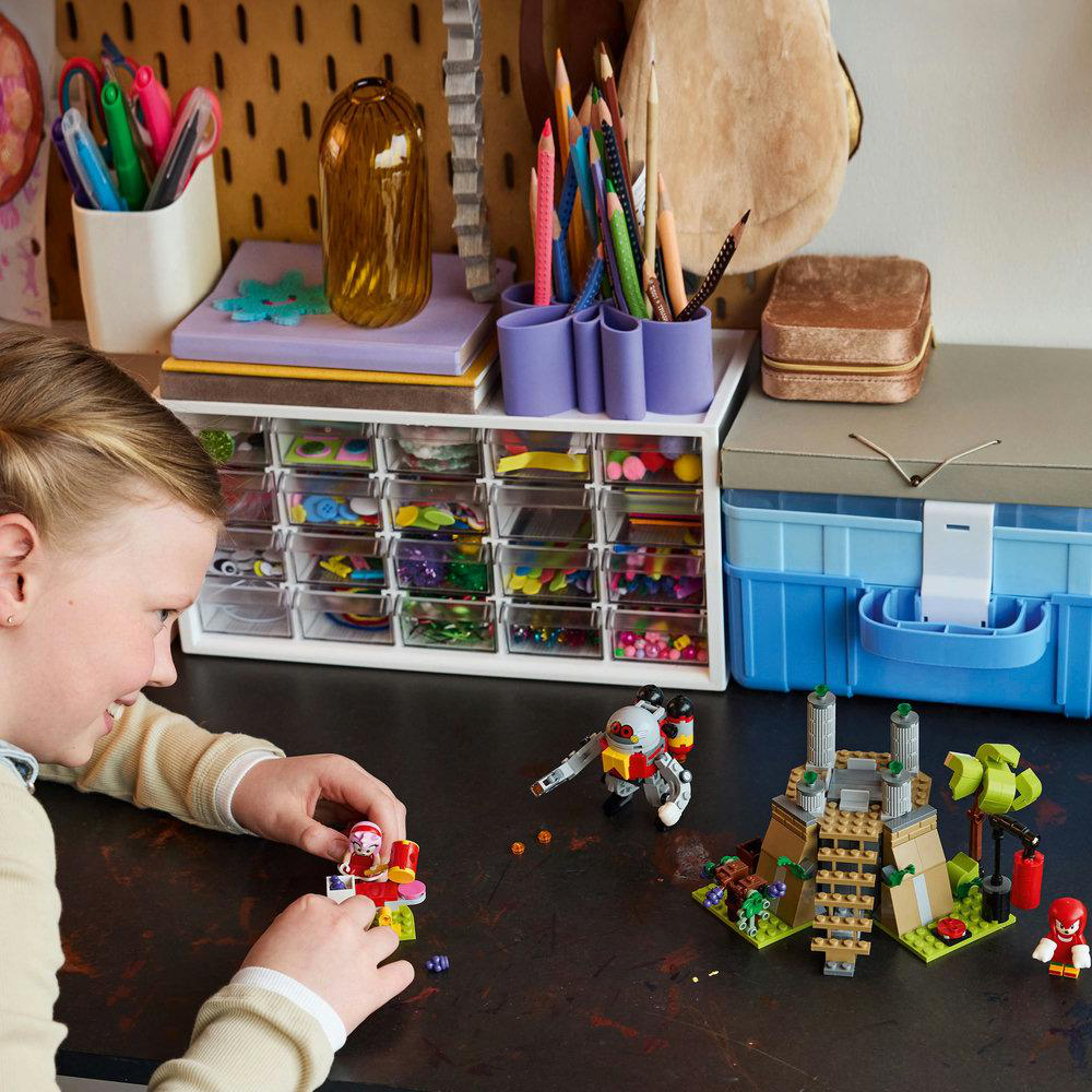 Mädchen spielt mit Lego-Steinen auf einem Schreibtisch, bunter Hintergrund mit Zubehör.