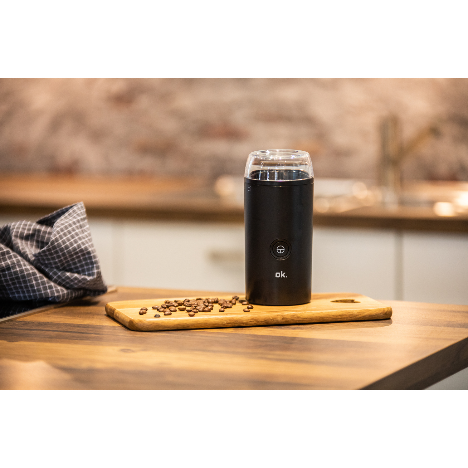 A black coffee grinder on a wooden board with coffee beans beside it. The background is blurred.
