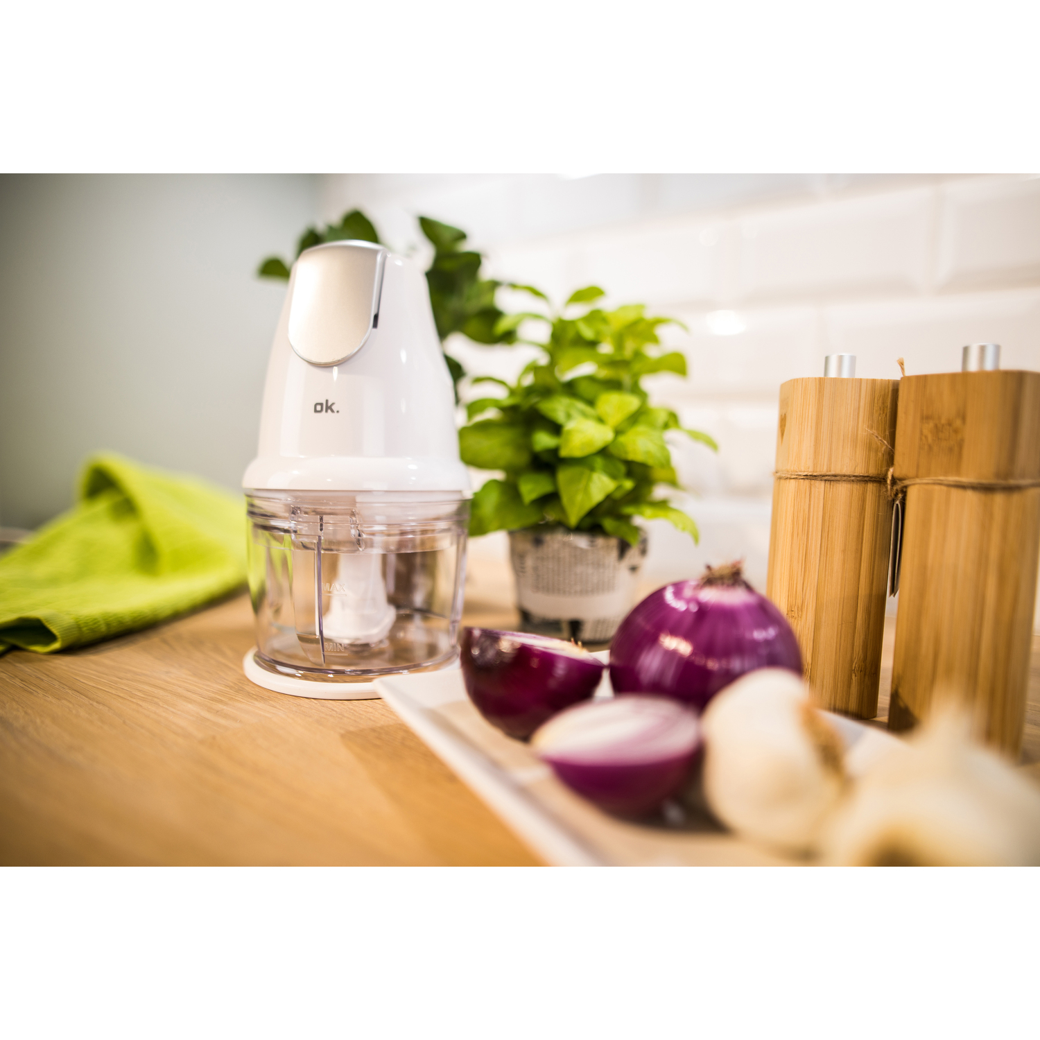 Een witte keukenmachine met uien, een plant en houten zout- en pepermolens op een tafel.