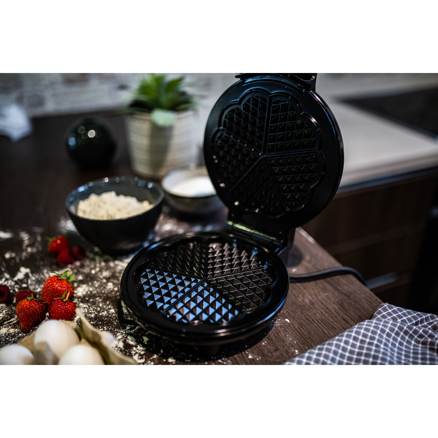 Gaufrier ouvert avec une surface noire. Ingrédients comme des baies et de la farine sont éparpillés.