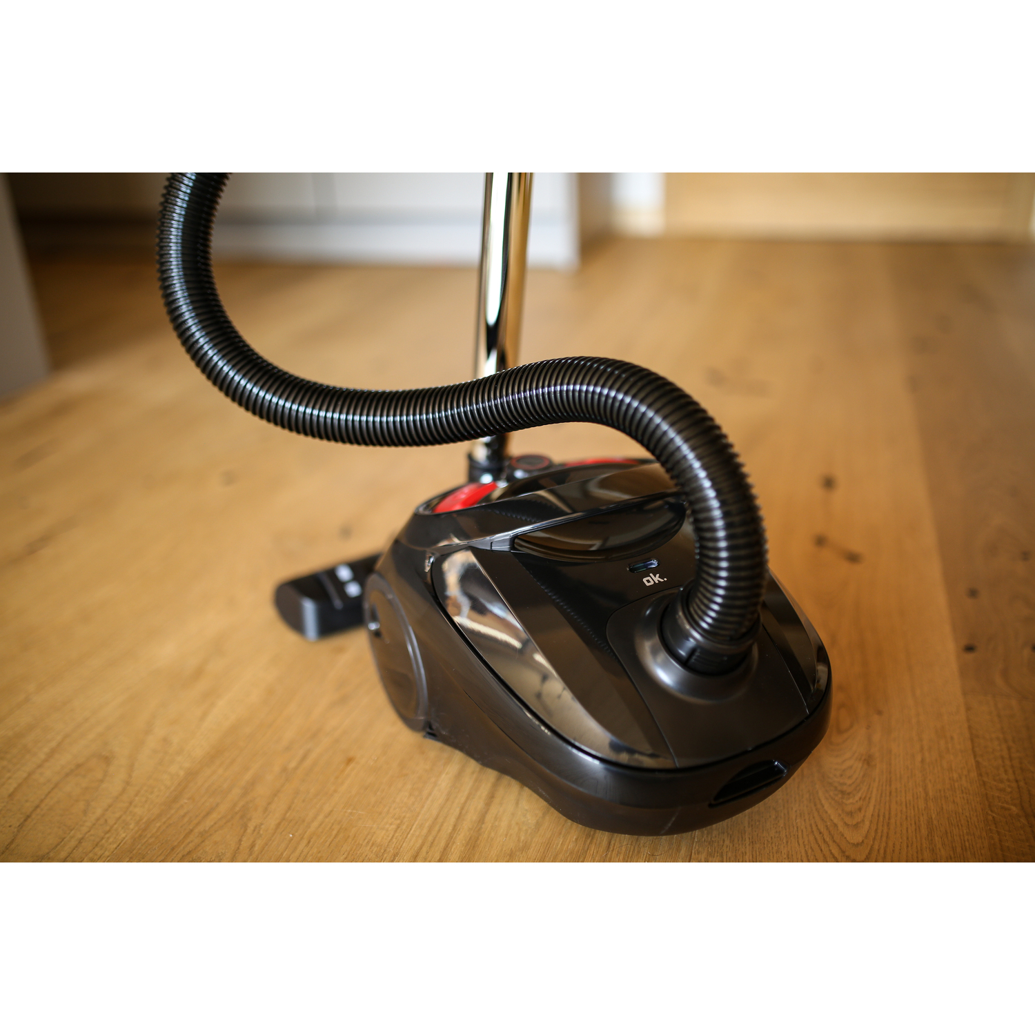 Black vacuum cleaner on wooden floor. The hose is curved. The floor is light brown.