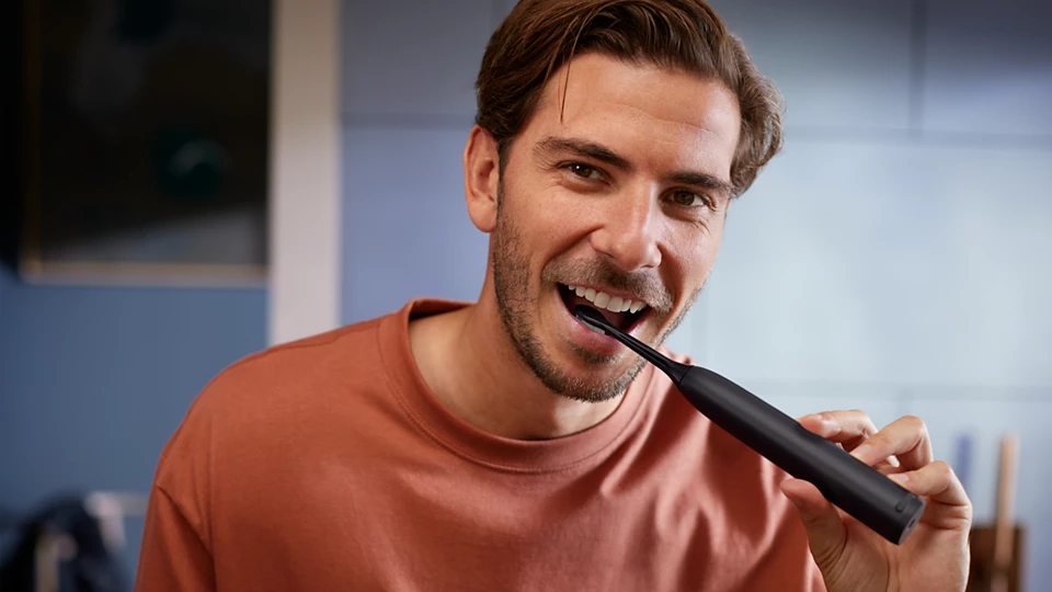 Un uomo si lava i denti con uno spazzolino elettrico nero. Sorride, indossa una maglietta arancione.