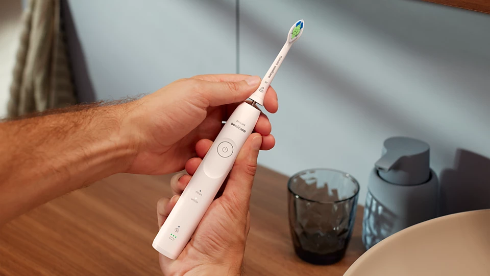 Mani che tengono uno spazzolino elettrico bianco. Un lavandino e un dosatore di sapone sullo sfondo.