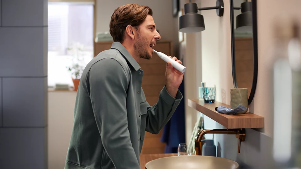 Uomo che si lava i denti in bagno, usando uno spazzolino elettrico, con riflesso nello specchio.