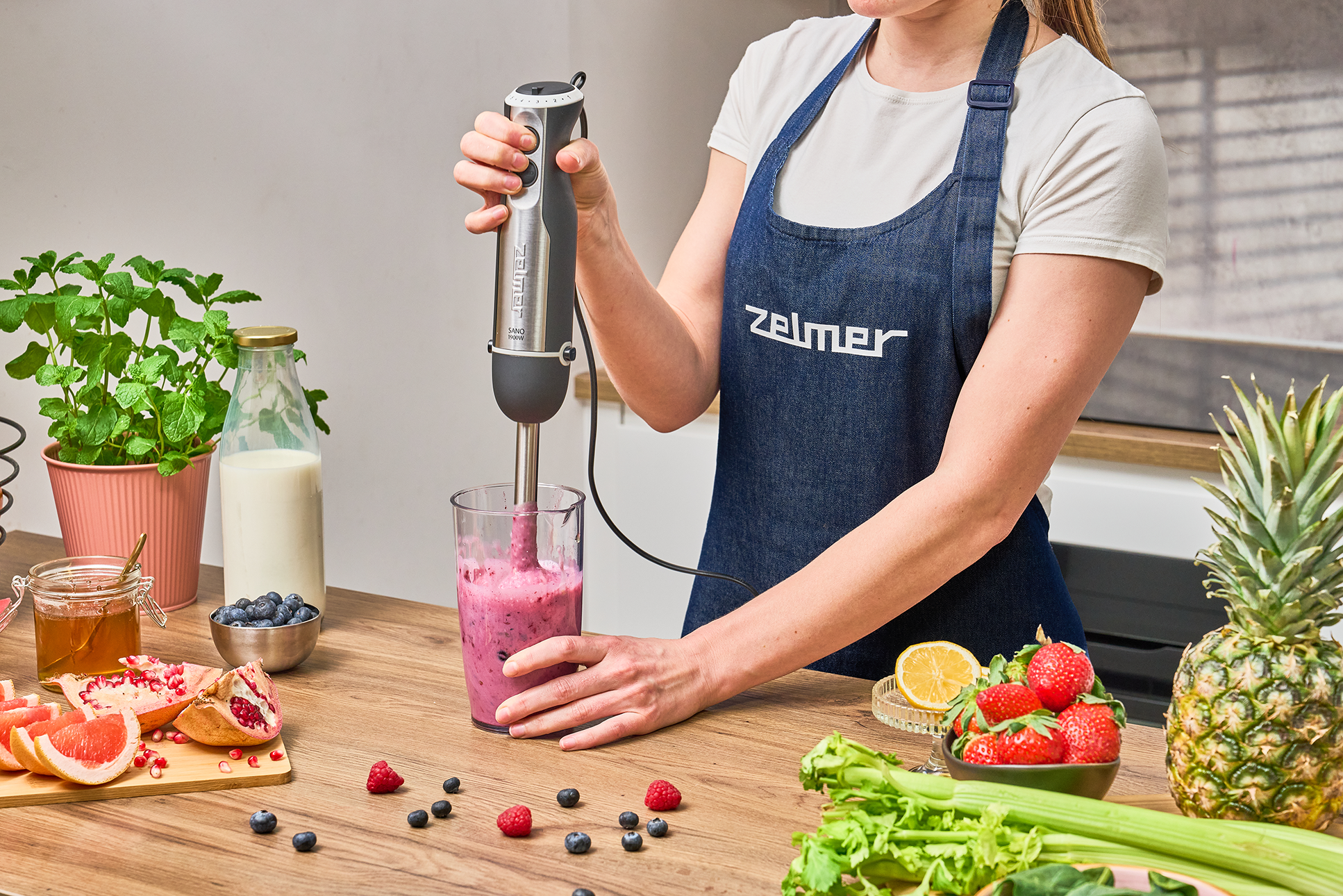 Egy személy turmixot készít botmixerrel, gyümölcsökkel és hozzávalókkal körülvéve.
