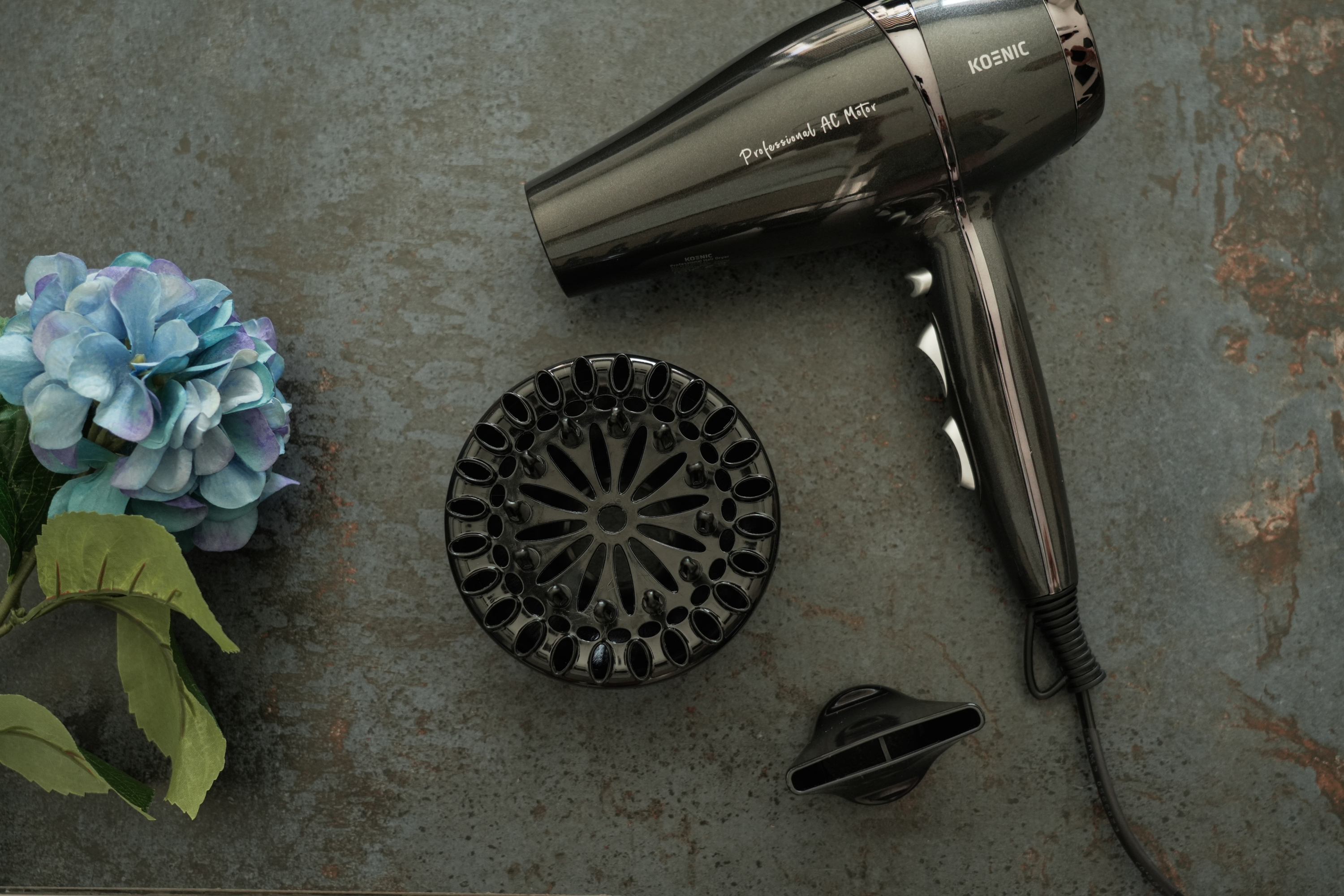 Czarna suszarka, dyfuzor i dysza na ciemnoszarej powierzchni z niebieskim kwiatem.