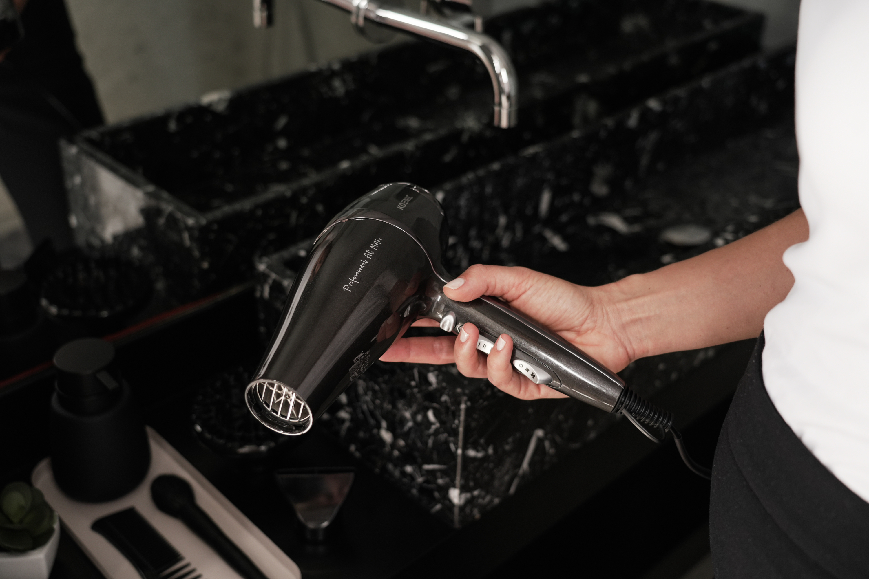 Osoba trzymająca czarną suszarkę do włosów, z lustrem i umywalką w tle.